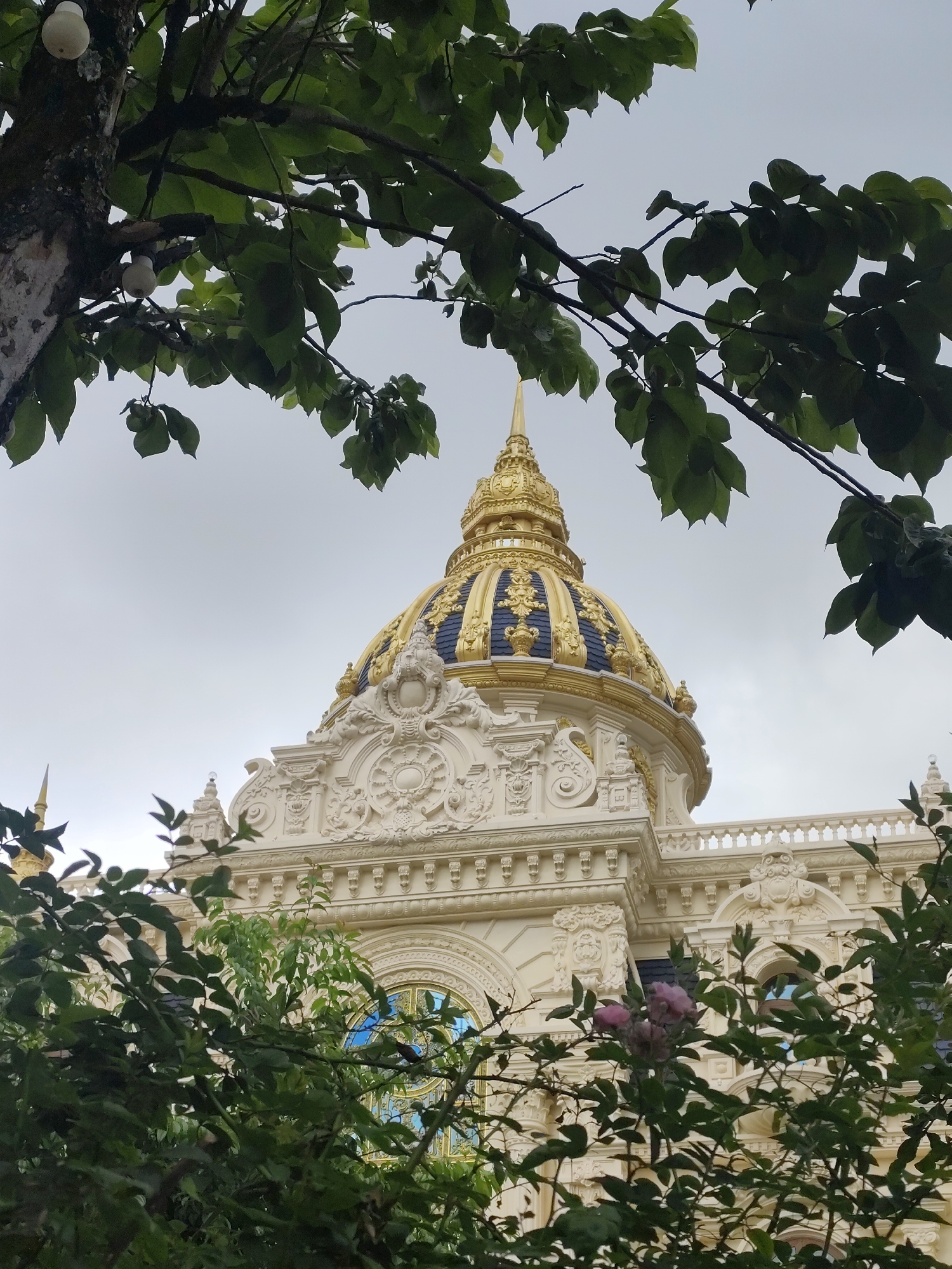 The image captures a portion of the Palace II in Da Lat, Vietnam, a historic French colonial building known for its distinctive architecture. In the foreground, lush green leaves and branches obscure parts of the view, creating a natural frame for the opulent structure. Dominating the scene is a golden dome, intricately decorated with dark blue accents and topped with a spire. The building itself is adorned with elaborate white stucco ornamentation, featuring swirling patterns, decorative reliefs, and arched windows. The sky above is overcast, with a pale, diffused light. Some round, white objects, possibly lights, are visible attached to the trunk of a tree on the left side of the image, partially obscured by leaves. A few pale pink flowers are also visible amidst the foliage. The overall impression is one of grandeur and historical significance, softened by the surrounding greenery.