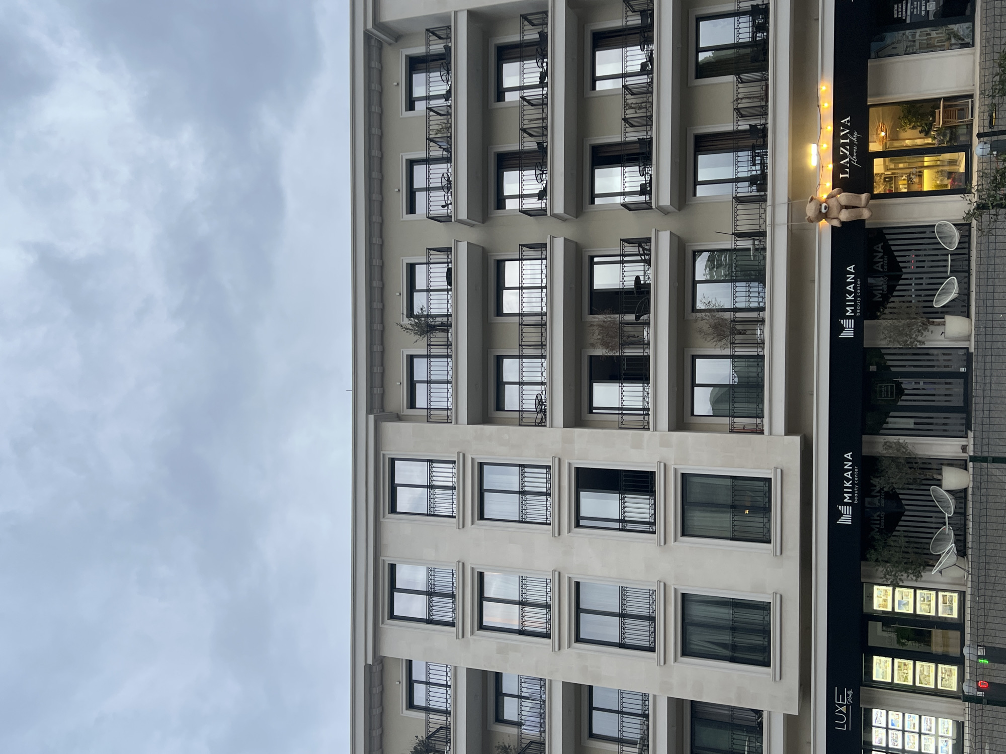 The image captures a building exterior, likely an apartment complex, under an overcast sky. The building features multiple floors with balconies, some adorned with plants and furniture. At ground level, business signs are visible, including "MIKANA beauty center," "LUXE REAL ESTATE," and "LAZIVA flower shop." A teddy bear is suspended above the entrance to "LAZIVA." The location is identified via other sources as Budva, Montenegro.
