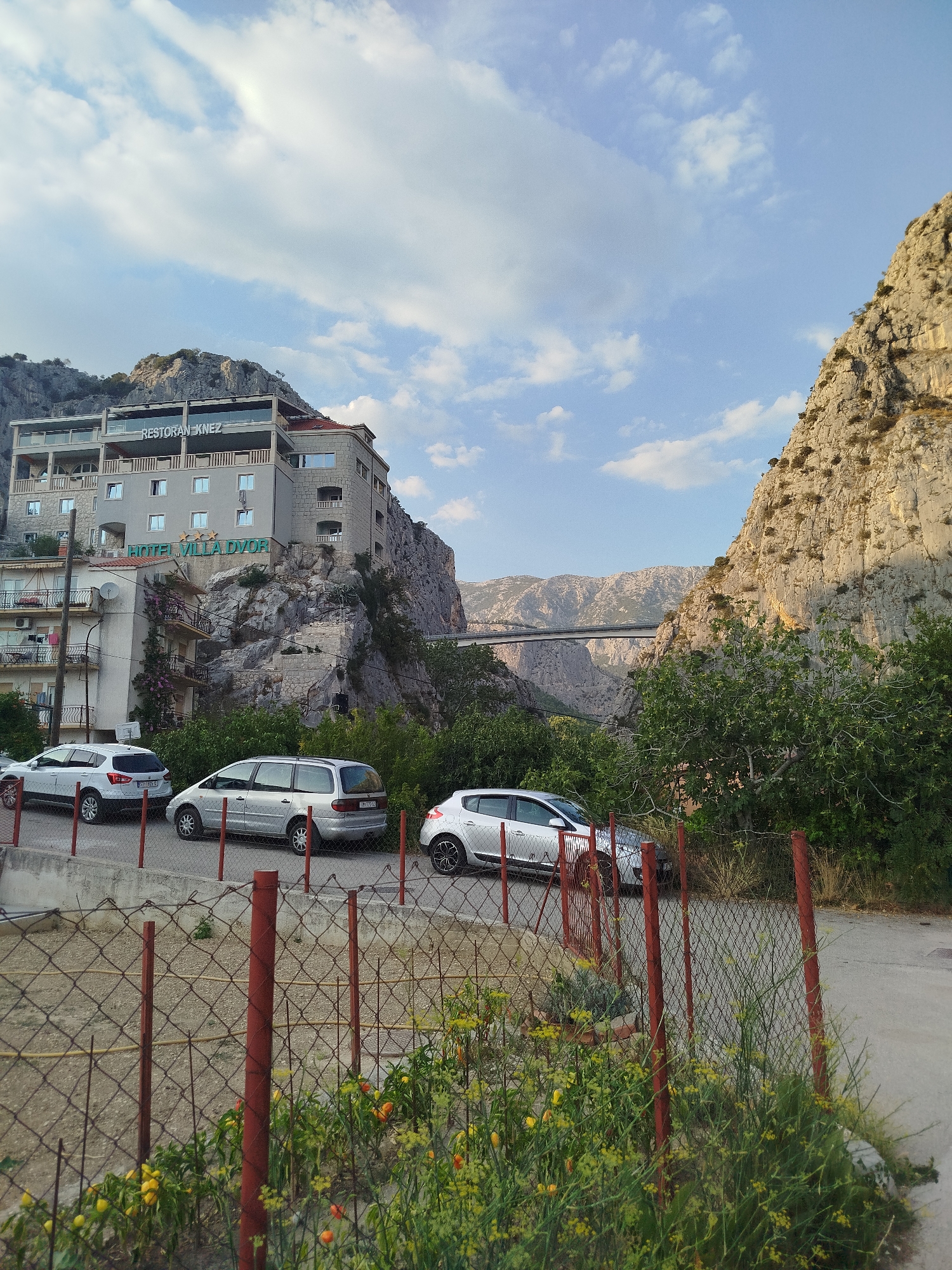 The image shows a view of the city of Omiš, Croatia, featuring a multi-story building with "HOTEL VILLA DVOR" and "RESTORAN KNEZ" signs, built into the side of a steep rocky cliff. Several cars are parked in front of the building, and a chain-link fence with red posts runs across the foreground, separating a garden with yellow and orange flowers from the parking area. In the background, a long bridge spans across a canyon, with more rocky hills and mountains visible in the distance under a partly cloudy sky. The scene appears to be captured during the late afternoon or early evening due to the lighting.