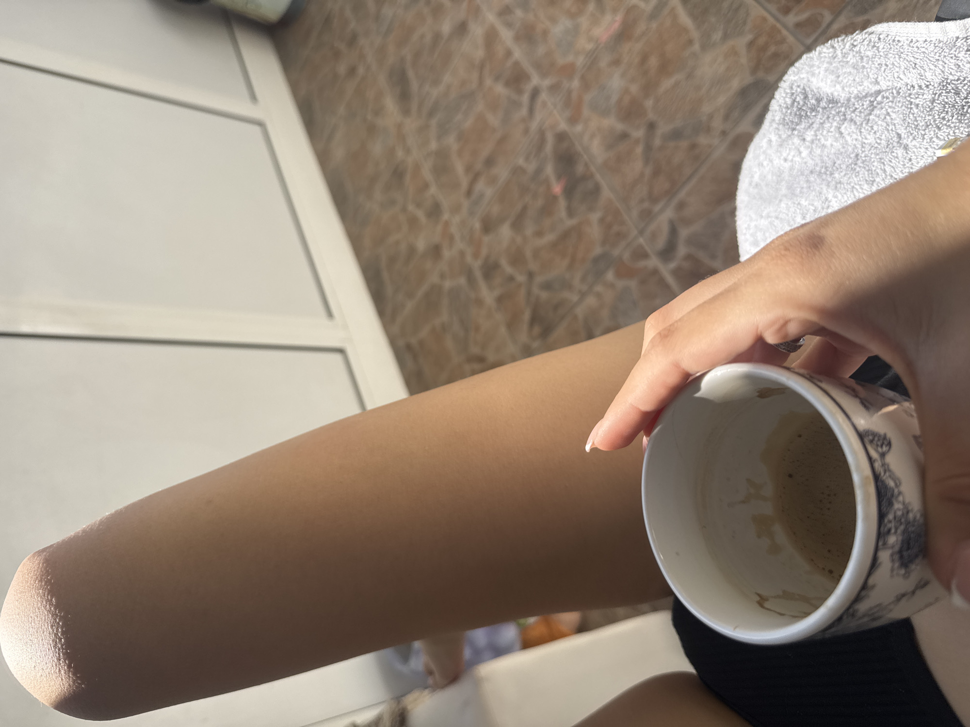 A person is holding a patterned ceramic mug containing the remnants of a dark liquid, likely coffee. The person's bare leg is in the foreground, positioned alongside a white door or window frame. The background features textured brown tiles and a white towel or fabric. The image was potentially taken outdoors, with natural light illuminating the scene. The location is unknown, but you requested the city of Plovdiv, Bulgaria.