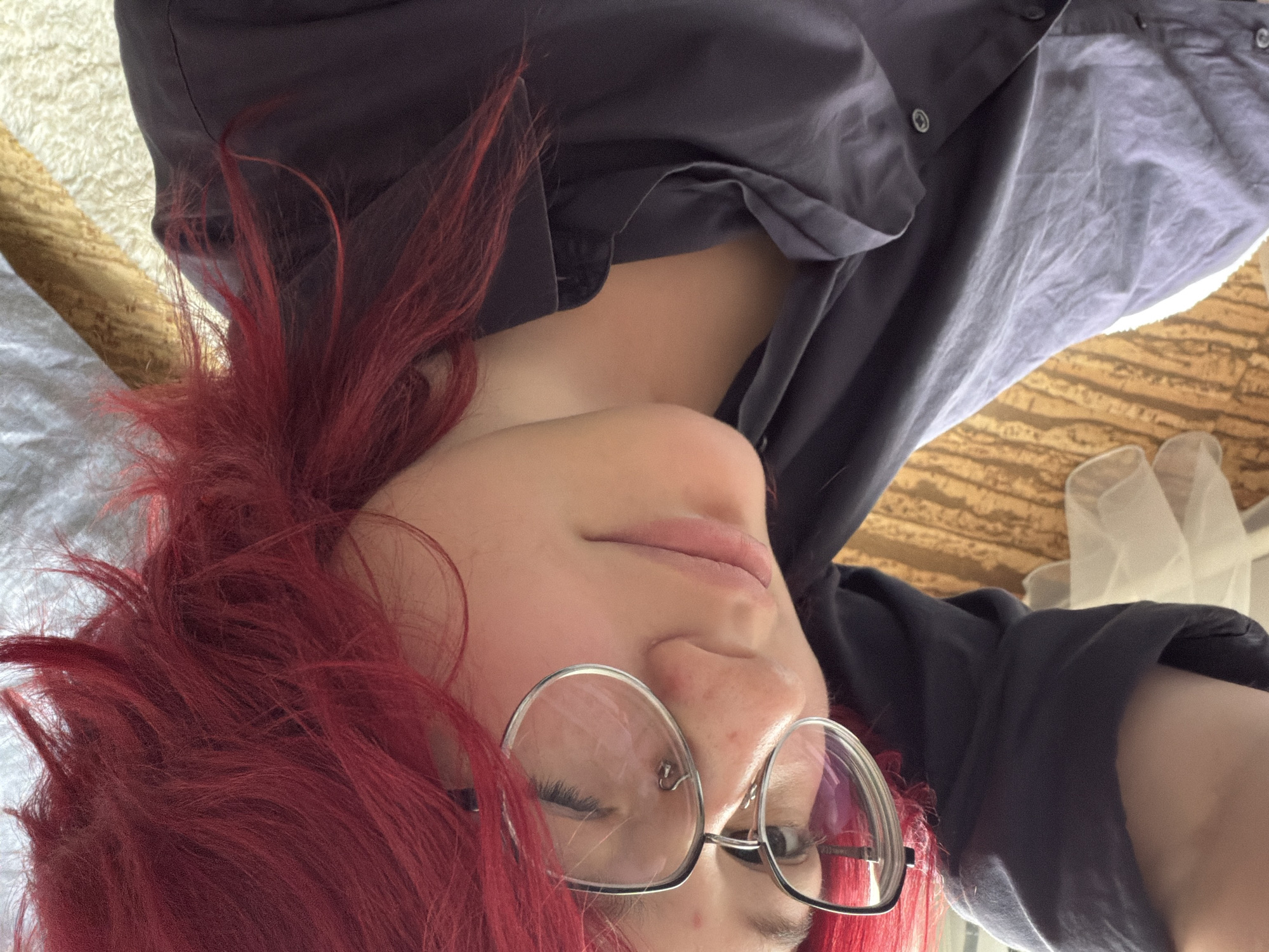 A person with bright red, spiky hair is lying down, looking up at the camera. They are wearing a dark grey button-up shirt, and large, round-rimmed glasses. The background is blurry and appears to be indoors, with a textured surface that could be a bedspread or carpet. There is no visible indication of the location within the city of Chelyabinsk, Russia.