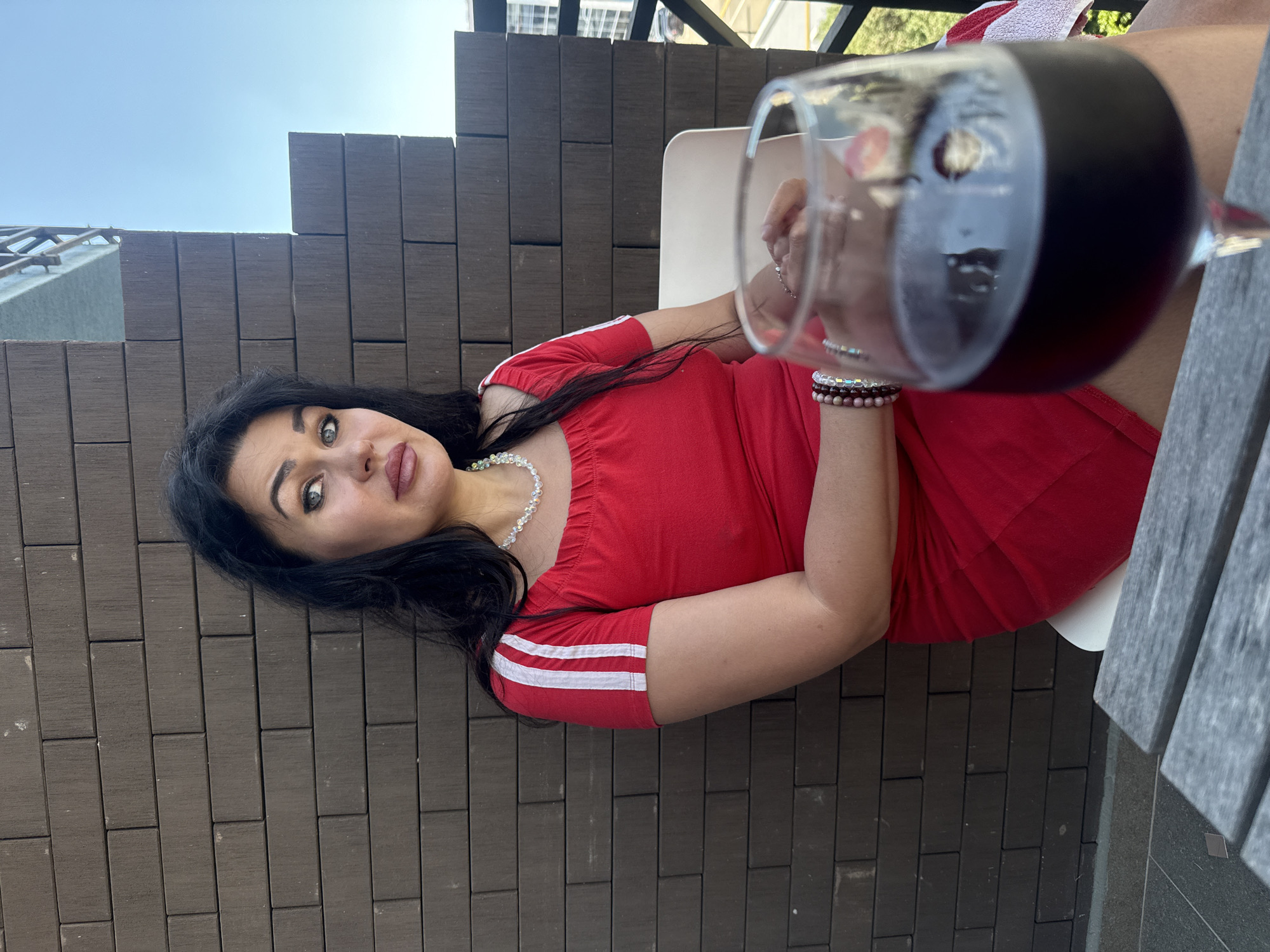 In Sochi, Russia, a woman with dark hair and green eyes is seated on a chair, wearing a red dress with white stripes on the sleeves. She has a beaded necklace and bracelets on her left arm. In the foreground, a glass of red wine is positioned on a table, with her hands resting on the table, partially obscured by the glass. The background features a brick wall and a clear blue sky.