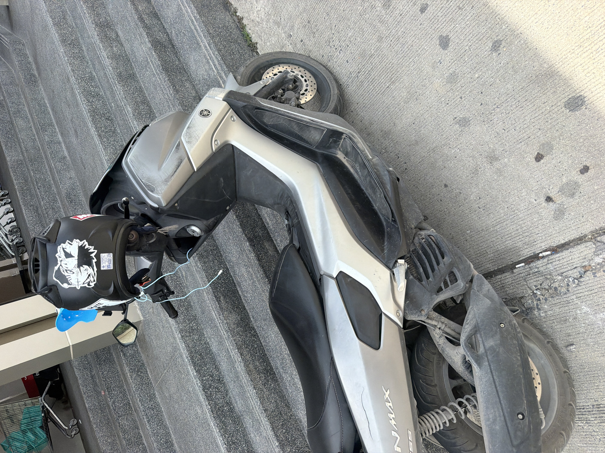 A silver Yamaha NMAX scooter is parked on a concrete sidewalk next to a set of grey speckled stairs. A black helmet with a sticker of an anime character is resting on the handlebars. A blue object, possibly a child's riding toy, is attached to the scooter's handlebars. The scooter appears to be slightly worn, with some scuff marks visible on its body and exhaust pipe. In the background, a shopping cart with teal items inside is visible on the stairs. The scene is set in an outdoor urban environment.