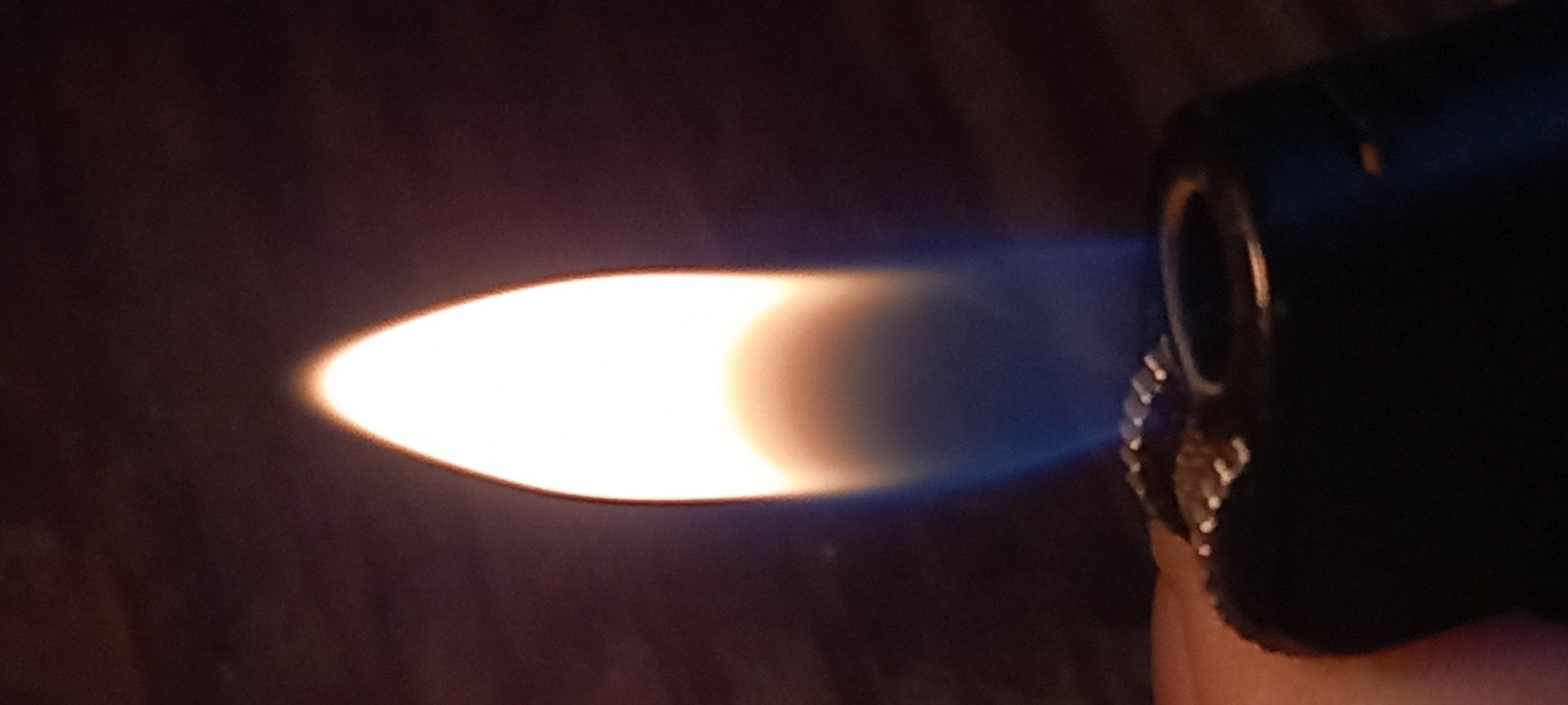 The image displays a close-up, horizontal view of a torch lighter actively producing a bright, powerful flame. On the right side of the frame, the dark body of the lighter is partially visible, along with a serrated ignition wheel and a glimpse of a finger or thumb holding it.
A prominent, elongated flame emanates from the lighter's nozzle, stretching diagonally across the center-left of the image. The flame is intensely bright at its core, appearing a luminous white and yellow, and transitions to a distinct, translucent blue as it extends outwards. This coloration indicates a high-temperature, clean-burning flame, typical of butane or torch lighters.
The background is dark, out of focus, and appears to be a muted brown and black, contrasting sharply with the brilliant light of the flame. This suggests the image was taken in low-light conditions, possibly indoors or at night. There are no other discernible objects or people, and no visible text in the image. The scene is located in Kryvyi Rih, Ukraine.