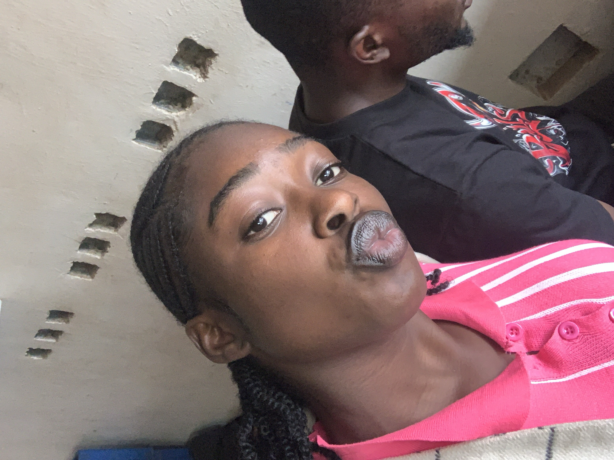 The image, captured in New Stadium, Nigeria, features two individuals in an indoor or semi-enclosed setting against a plain, light-colored wall. In the foreground, a young woman with dark skin and dark braided hair is prominently displayed, looking directly at the camera with her lips pursed. She wears a pink, vertically ribbed top with white stripes and a button-down placket, partially obscured by a light-toned, possibly striped, garment draped over her left shoulder. To her right and partially obscured, a man with dark skin and a beard is visible from the neck up and across his upper torso. He is wearing a dark, short-sleeved t-shirt with a red and white graphic design on the chest and is looking downwards, away from the camera. The background wall exhibits multiple small, roughly rectangular indentations or holes, scattered irregularly across its surface. A blurred blue object is discernible in the upper left corner of the frame.