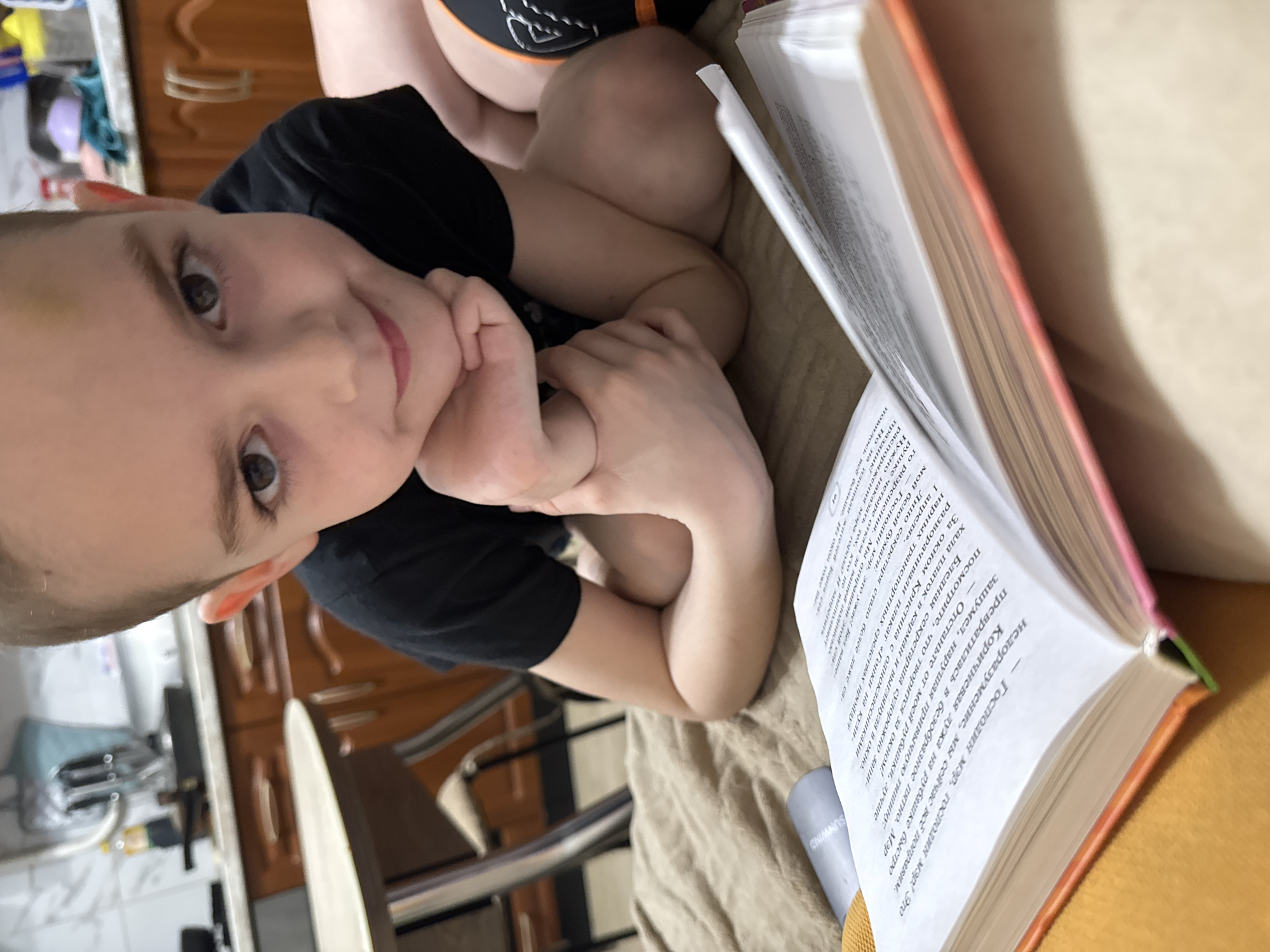 The image, rotated approximately 90 degrees counter-clockwise, features a young child looking directly at the camera with a calm expression, resting their chin on their hands. The child is wearing a black t-shirt. To the right, an open book with visible text is presented, held by an individual whose legs and upper torso are partially visible, suggesting they are also part of the reading activity.
The scene is set indoors, likely in a home in Razdolye, Russia. In the background, a kitchen area with wooden cabinets, countertops, and a bottle (possibly cooking oil) can be seen. A chair is also visible. The lighting suggests it is daytime. The overall atmosphere appears peaceful and domestic.
Notable visible text from the open book is in Russian, describing a dramatic narrative: "Mr. Mayor, Mr. Mayor! This is a misunderstanding, we will fix everything now." and "Get off my shirt, better look what's happening outside the window!". Other phrases mention "Kristina with Classmates," a "poster 'Races on stilts- April'," and "The secretary's face turned even whiter...". A device or speaker in the foreground has the brand name "SUNWIND" printed on it.