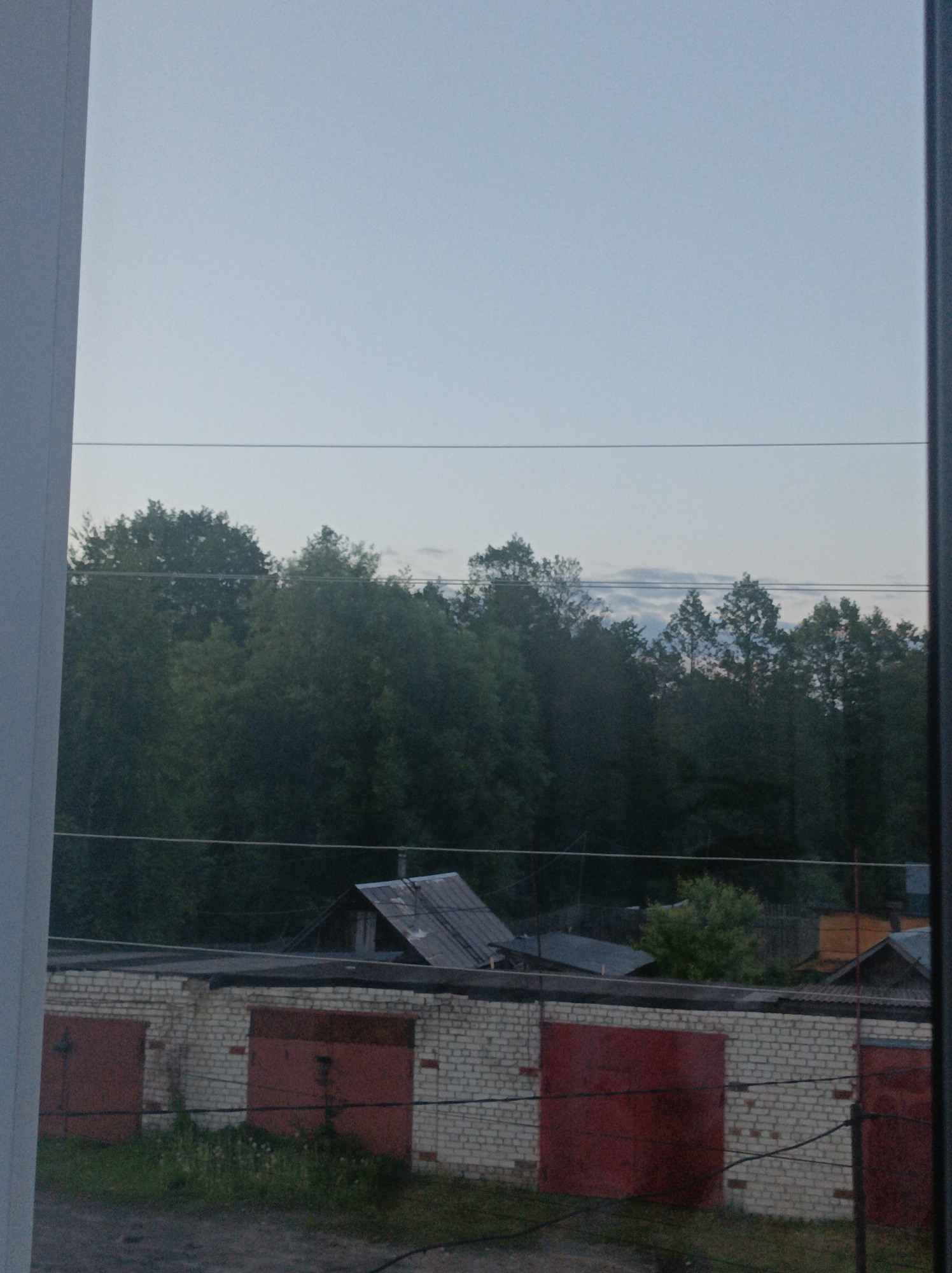 The image captures an outdoor scene, likely viewed through a window due to the vertical white and dark grey frames on the left and right, respectively. The upper third of the frame is dominated by a pale, hazy blue-grey sky, suggesting either dusk, dawn, or an overcast day. Faint clouds are visible towards the horizon.
Below the sky, a dense band of dark green trees stretches across the middle ground, forming a natural barrier. In front of the treeline, a row of single-story garages is prominent. These garages are constructed from light-colored, possibly white or cream, brick and feature multiple weathered, reddish-brown metal doors, some showing visible rust. The ground in front of the garages is a mix of dirt or gravel with sparse green grass and weeds.
Behind the garages and partially obscured by the trees, a small, rustic building with a steep, corrugated metal roof is visible. Other faint structures or roofs can be seen further in the background to the right. Several thin, dark horizontal lines, likely power or utility wires, traverse the image at various levels. No people or discernible activities are present. There is no visible text. The overall setting suggests a quiet, possibly semi-rural or industrial fringe area in Semyonov, Russia.