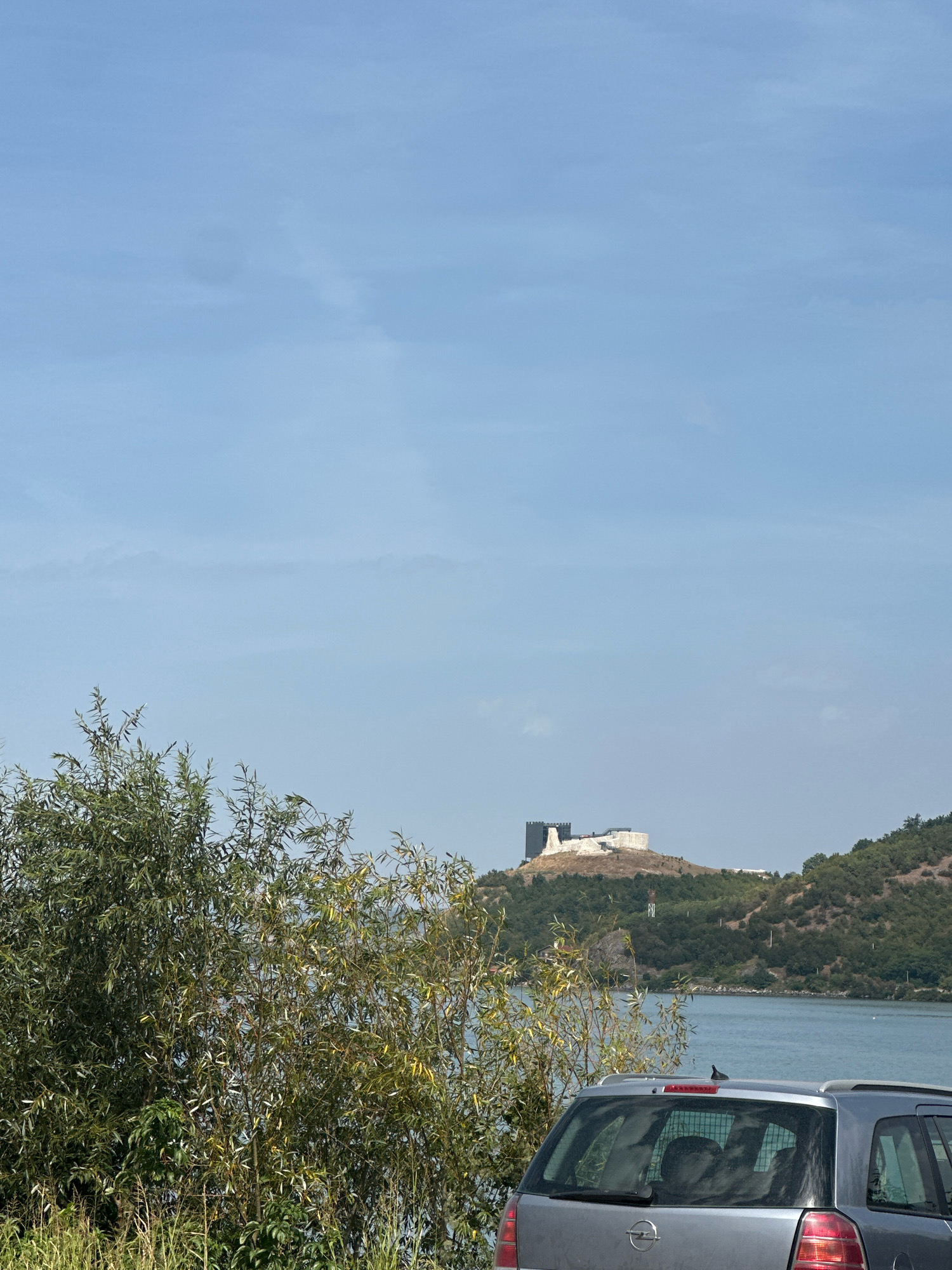 The image shows a view of the Golubac Fortress, a medieval castle situated in the city of Golubac Municipality, Serbia. The fortress is perched atop a hill overlooking a body of water, likely the Danube River. In the foreground, a silver Opel hatchback car is parked, partially obscuring a lush green bush with a mix of foliage and dry grasses. The sky above is a pale blue with sparse clouds. The fortress itself is a prominent structure with stone walls and towers, some of which appear to be undergoing renovation or are partially in ruins. The surrounding landscape is characterized by rolling green hills covered in trees. The scene suggests a clear day with a relaxed atmosphere, possibly a tourist stop. There are no people visible in the image.