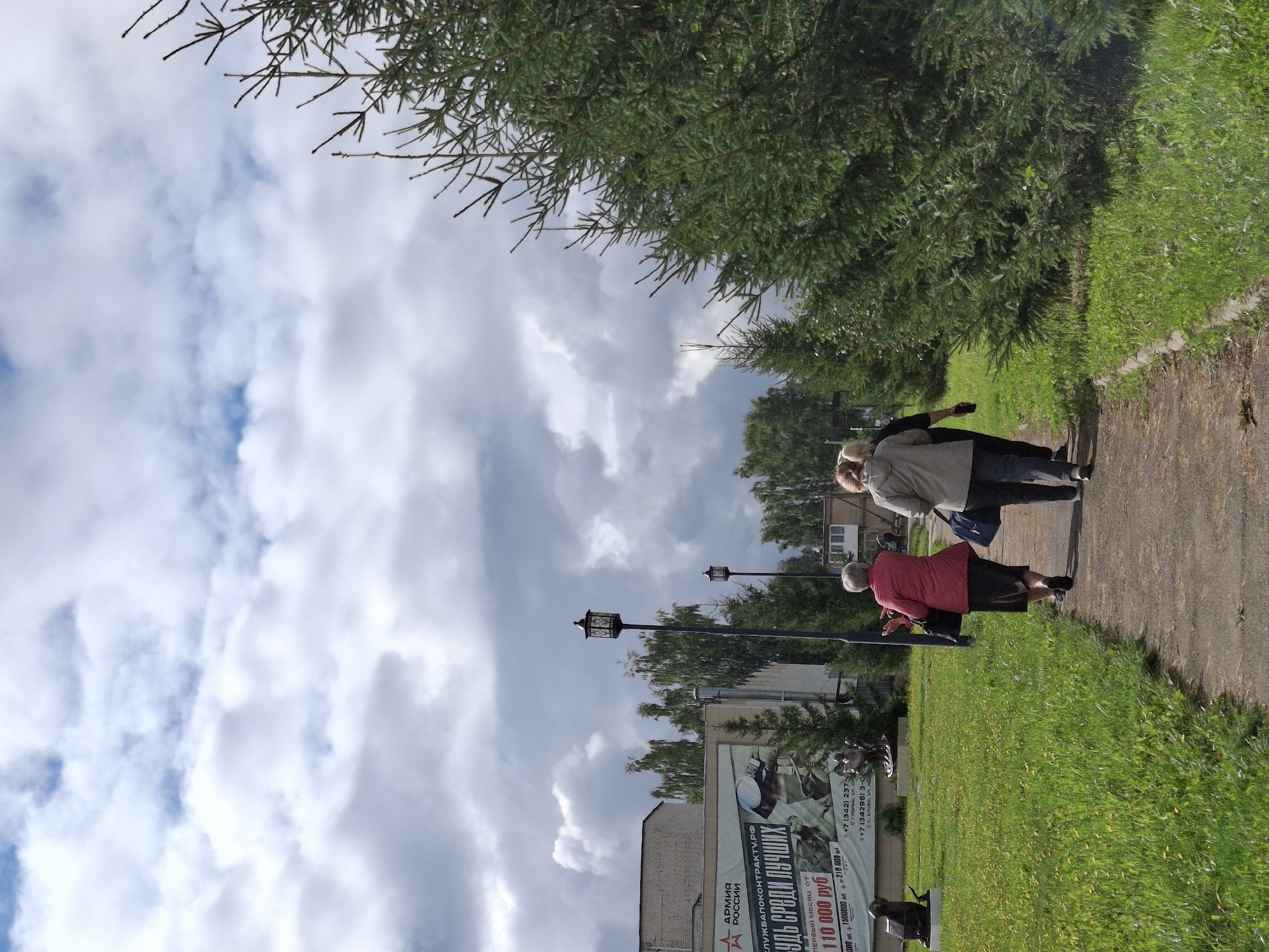 In Elovo, Russia, three individuals are walking away from the camera on a paved path bordered by green grass. The person in the foreground, wearing a pink jacket and dark skirt, is walking ahead of two others. One of them wears a grey hooded jacket and dark pants, carrying a blue bag. The third individual, to the far right, wears a light grey jacket and dark pants, also carrying a blue bag. To the left of the path is a grassy area with some yellow wildflowers and a black, angular sculpture resembling an animal. Further left, a large billboard displays military recruitment information with Russian text, an image of a soldier, and contact details including a phone number and website. Behind the billboard, a multi-story building stands. Two lampposts with decorative black lanterns are visible on the path, and behind them, trees line the horizon. A dense evergreen tree on the right side of the image partially obscures the view. The sky above is filled with a mix of bright white and darker grey clouds.