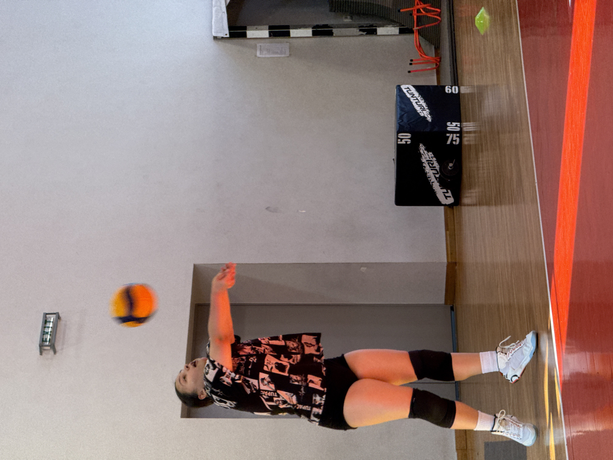 A woman is actively engaged in playing volleyball indoors. She is wearing a shirt with black and white graphics, black shorts, knee pads, white socks, and light blue tennis shoes. A volleyball, featuring a mix of blue and orange colors, is suspended in the air near her left hand.
The scene appears to be inside a gymnasium or sports hall in Etropole, Bulgaria. The background includes a light grey wall with a silver fixture and a doorway. On the floor to the right, there is a black mat with numbers printed on it, ranging from 50 to 75, adjacent to a red strip along the edge of the court. A yellow object is suspended from the ceiling. The image is rotated 90 degrees clockwise.