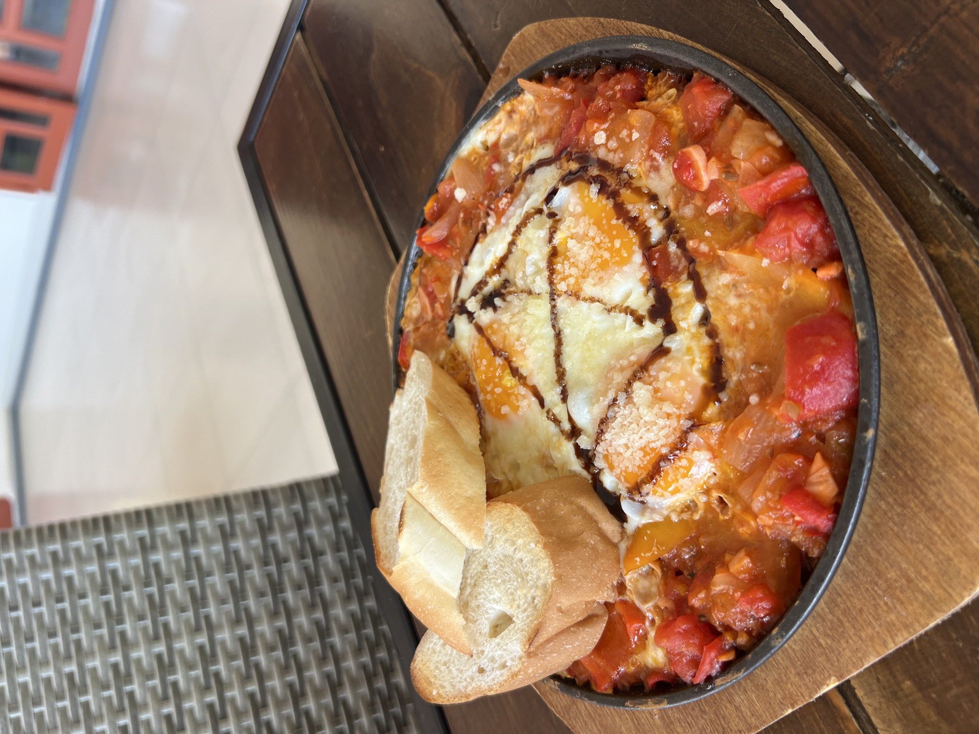This overhead shot captures a vibrant and appetizing meal served on a wooden table, likely in an outdoor or semi-outdoor dining setting during the daytime in Bac Nha Trang Ward, Vietnam.
The main subject is a round, black cast-iron pan, possibly still sizzling, containing a dish resembling Shakshuka or a similar egg and tomato-based preparation. The pan is filled with a rich, reddish-orange sauce, abundant with chunky diced red tomatoes, pieces of what appear to be yellow and red bell peppers, and possibly onions. Nestled within this savory base are at least two perfectly cooked eggs, with visible whites and bright yellow yolks, some of which are still runny. The entire dish is garnished with a generous sprinkle of grated white cheese, resembling Parmesan, and a decorative zigzag drizzle of a dark, glossy sauce, possibly balsamic glaze.
Accompanying the main dish are several slices of light-colored, crusty baguette bread, placed neatly next to the pan, presumably for dipping. The pan itself rests on a dark, rustic wooden serving board or trivet. The table beneath is made of dark wood planks. In the background, to the left, a light-colored surface, possibly a floor or wall, is visible, along with a section of a gray, woven-patterned surface, suggesting a chair or other outdoor furniture. The bright, natural illumination indicates that the photograph was taken during daylight hours. No visible text is present in the image.
