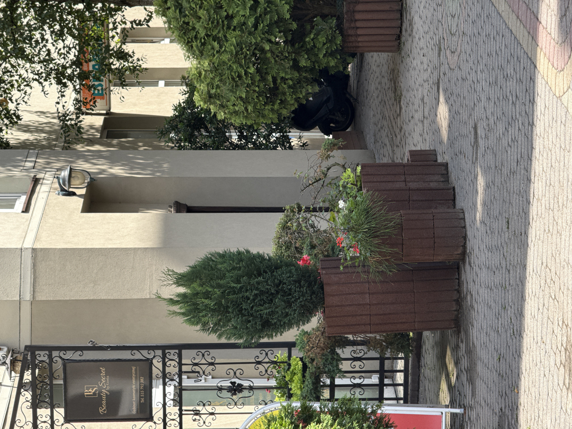 In Łódź, Poland, outside a building with a tan facade, a black wrought iron gate leads to an entrance. A sign on the gate reads "Beauty Secret by Gosia." Several large, cylindrical planters made of brown concrete blocks hold various green plants, including a tall conical evergreen in the foreground. Red flowers are visible in some of the planters. A black scooter is parked behind a large green shrub to the right. The ground is paved with interlocking gray bricks. A portion of a building with windows and an outdoor lamp is visible above the gate. Sunlight casts shadows on the pavement.