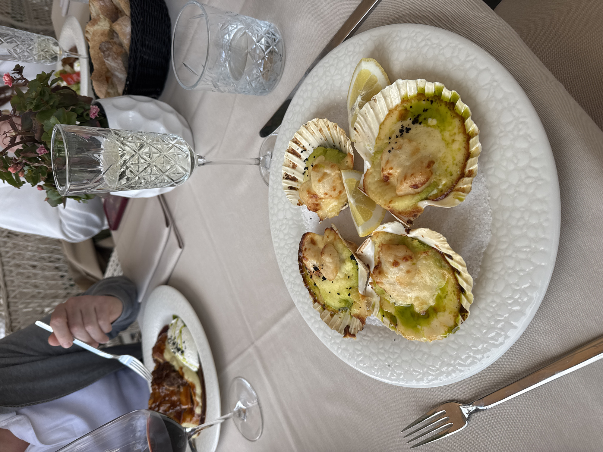The image shows a table setting at a restaurant, likely in Radovići, Montenegro, as specified. There are two diners visible, one partially shown with a fork in hand. The table is covered with a light beige tablecloth. The main focus is a white plate with four scallops served in their shells, garnished with lemon wedges and sprinkled with black seeds. There is a second plate with food on the left side. The table setting includes crystal glasses for water and wine, along with silverware. A basket of bread and a small floral arrangement are placed near the top left corner. The overall ambiance suggests a restaurant setting.