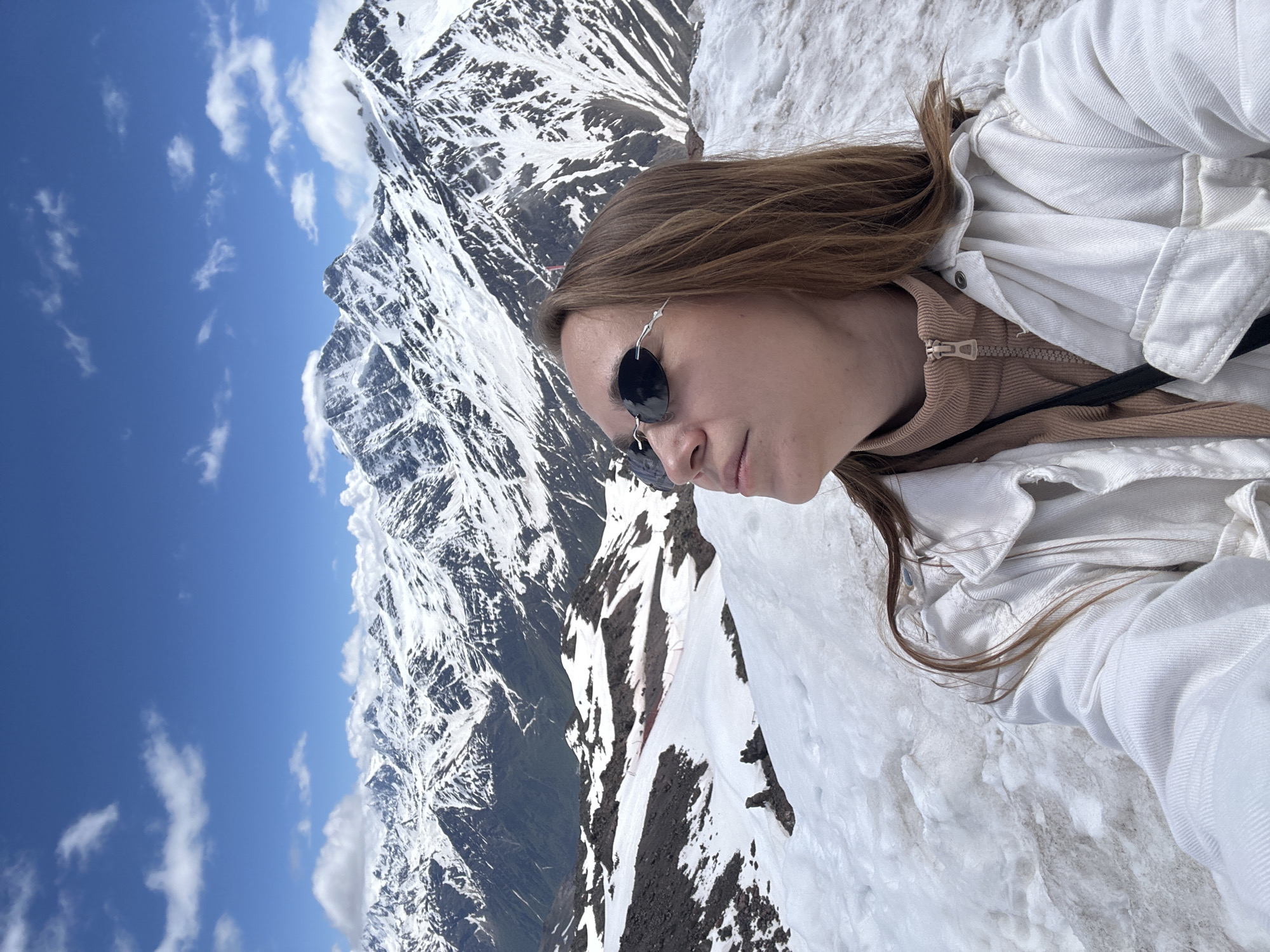 A medium shot captures a woman with long, brown hair wearing sunglasses, a tan turtleneck, and a white jacket. She is looking upwards and to the left, her head angled slightly. The background is a majestic mountain landscape covered in snow, with a clear blue sky dotted with white clouds. The scene appears to be in the vicinity of Terskol, Russia, known for its mountainous terrain and ski resorts. The woman is positioned in the foreground, with the snow-covered mountains stretching out behind her under a bright, sunny sky.