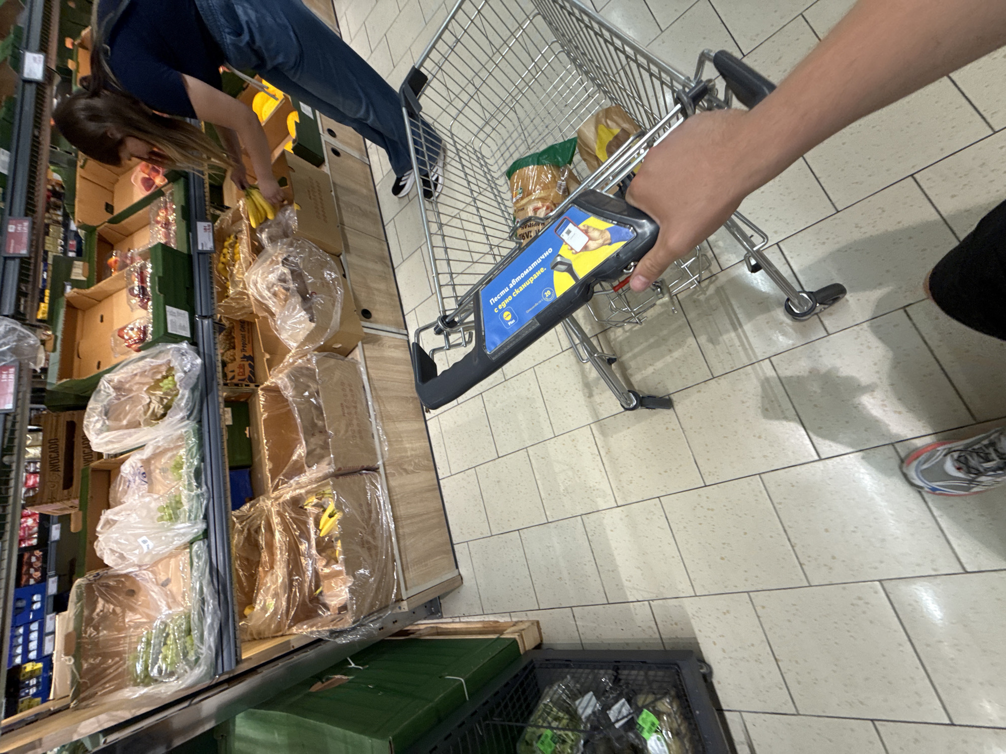 The image shows a first-person perspective of someone pushing a shopping cart in a supermarket. The cart has a blue sign with Ukrainian text and an illustration of a smartphone scanning a barcode. Inside the cart, there are several packaged items. In the background, a woman with long brown hair wearing a dark blue t-shirt and blue jeans is selecting bananas from a display. The shelves behind her are stocked with various fruits and vegetables, including grapes and avocados. The floor is tiled with large, light-colored tiles. The overall environment suggests a retail setting, likely a supermarket aisle. There is no indication that the location is specifically Stara Zagora, Bulgaria.