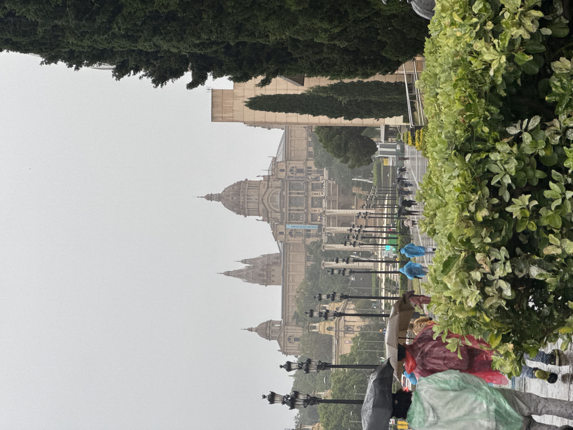 The image shows the Palau Nacional in Barcelona, Spain, seen from a lower elevation on a rainy day. The sky is overcast, contributing to the dull lighting.
People are visible on the pathway, several wearing colorful raincoats, and some holding umbrellas. The pathway is lined with ornate lampposts. The Palau Nacional is the most prominent structure, with its multiple domes and detailed facade. A few trees appear in the background as well as framing the sides of the image.