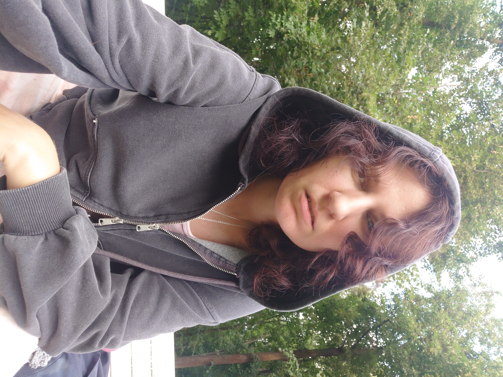 A woman with curly brown hair wears a dark gray hooded sweatshirt and a pink undershirt. She is seated outdoors with a background of green trees. Her expression is neutral, and she is looking directly at the camera. The lighting suggests it is daytime.