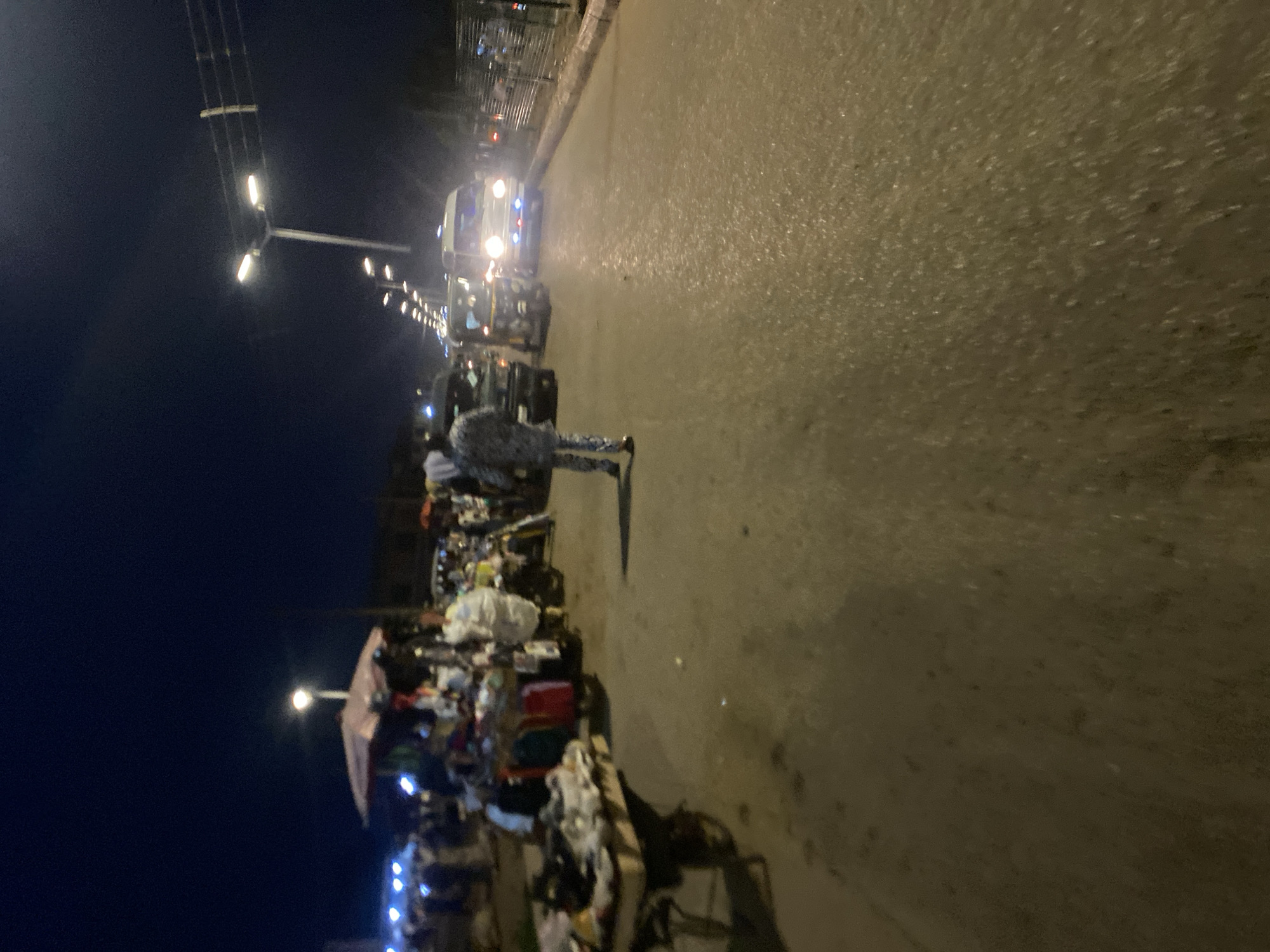 The image depicts a nighttime street scene in Bukuru, Nigeria. A person in a patterned outfit walks away from the camera on a wet asphalt road. To the left, market stalls with various goods are set up, including clothing and other items, with an umbrella overhead. Several vehicles, including a white van with flashing blue lights and a three-wheeled auto-rickshaw, are present on the road. Streetlights illuminate the scene, casting a yellowish glow. The sky is dark, suggesting late evening or night. The overall impression is of a busy, active marketplace at night.