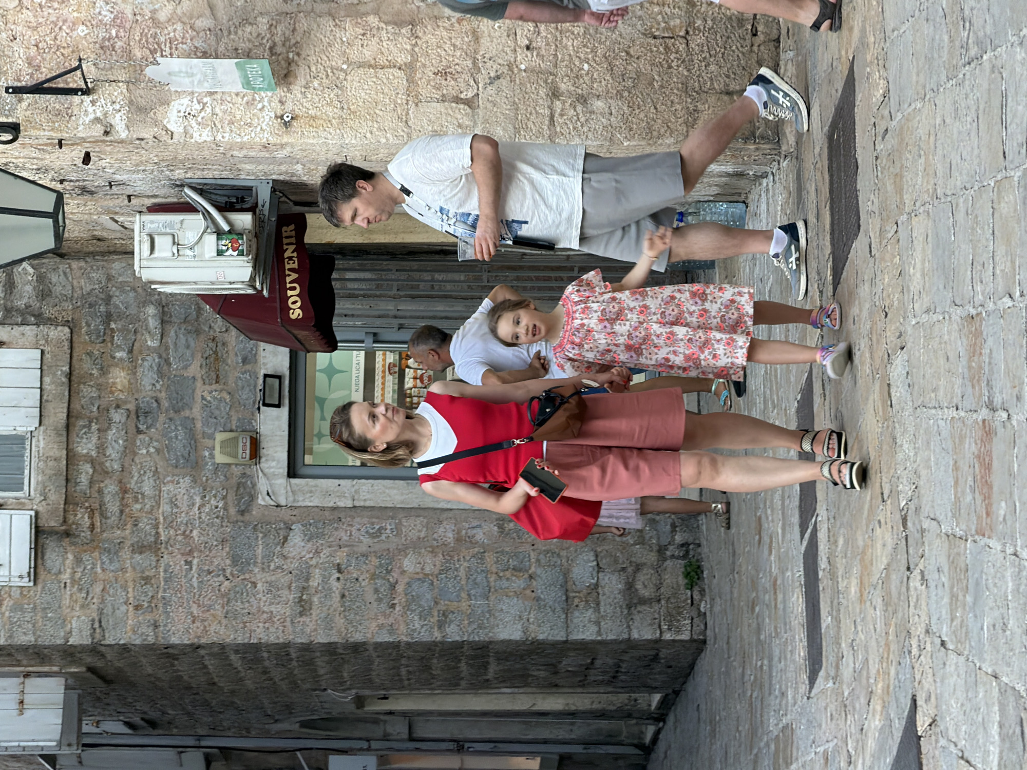 A family walks down a cobblestone street in the old town of Budva, Montenegro. The father, wearing a white t-shirt and grey shorts, holds a drink in his right hand and has a lanyard around his neck. Beside him, his daughter, in a floral print dress, walks with her left hand outstretched. The mother, in a red tank top and pink shorts, holds a red phone in her left hand and carries a brown crossbody bag. Another child in a pink dress is partially visible behind her. The street is lined with stone buildings, one of which has a "SOUVENIR" awning. An air conditioning unit is mounted on the exterior wall of one building.