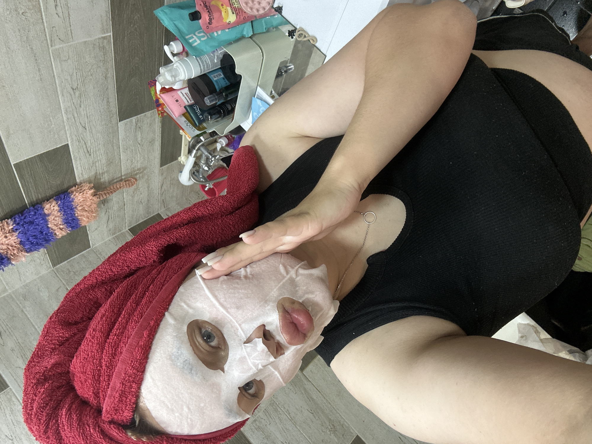 An individual is depicted indoors, presumably in a bathroom, engaged in a self-care routine. The person, appearing to be female, is wearing a white sheet face mask that covers the majority of the face, with cutouts for the eyes, nose, and mouth. Visible features include light-colored eyes and full, glossy pink lips. Her hair is wrapped in a red terry cloth towel, styled as a turban. She is wearing a black ribbed tank top and a thin silver necklace with a circular pendant. Her left hand, featuring long, light-colored fingernails, is raised with fingers splayed, resting lightly against the side of her masked face.
The background shows a tiled wall with a pattern of alternating light grey/beige and darker grey rectangular tiles, arranged horizontally in a staggered brick layout. To the right, on a counter or shelf, an assortment of beauty and skincare products is organized, some within a light-colored caddy. Visible brand names include "SADOER," "MIGIT," "LIFTING," and "ORGANIC KITCHEN," along with various bottles, tubes, and pumps. A chrome faucet and shower fixture are visible on the left side of the counter. A multi-colored knitted or crocheted object, featuring purple, light blue, and pink/peach stripes with a tassel-like end, hangs on the wall in the upper left portion of the image. No identifying markers indicating a specific city or country, such as Chaikovsky, Russia, are discernible within the image.