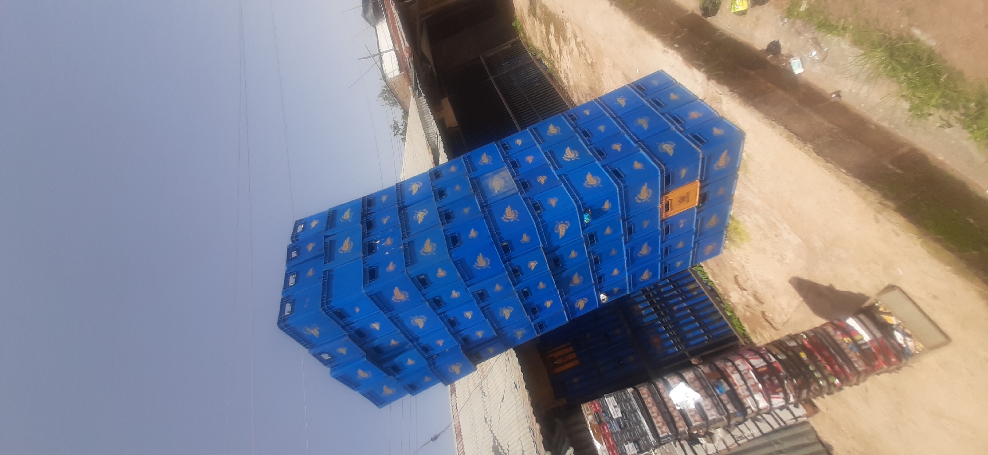 In the city of Jos, Nigeria, a very tall stack of blue plastic crates, possibly for beverages, is precariously balanced outdoors. The crates are uniformly blue and have a golden emblem on their sides, resembling a bird or a stylized leaf. The stack leans significantly to the left, creating a dramatic visual. In the background, there are low-rise buildings with corrugated metal roofs and some fencing. To the left of the main stack of crates, there is another, smaller stack of various items, including what appear to be packaged goods or boxes. The ground is a mixture of dirt and concrete, with some patches of dry grass. The sky above is a clear, pale blue. The scene appears to be an outdoor storage or market area.