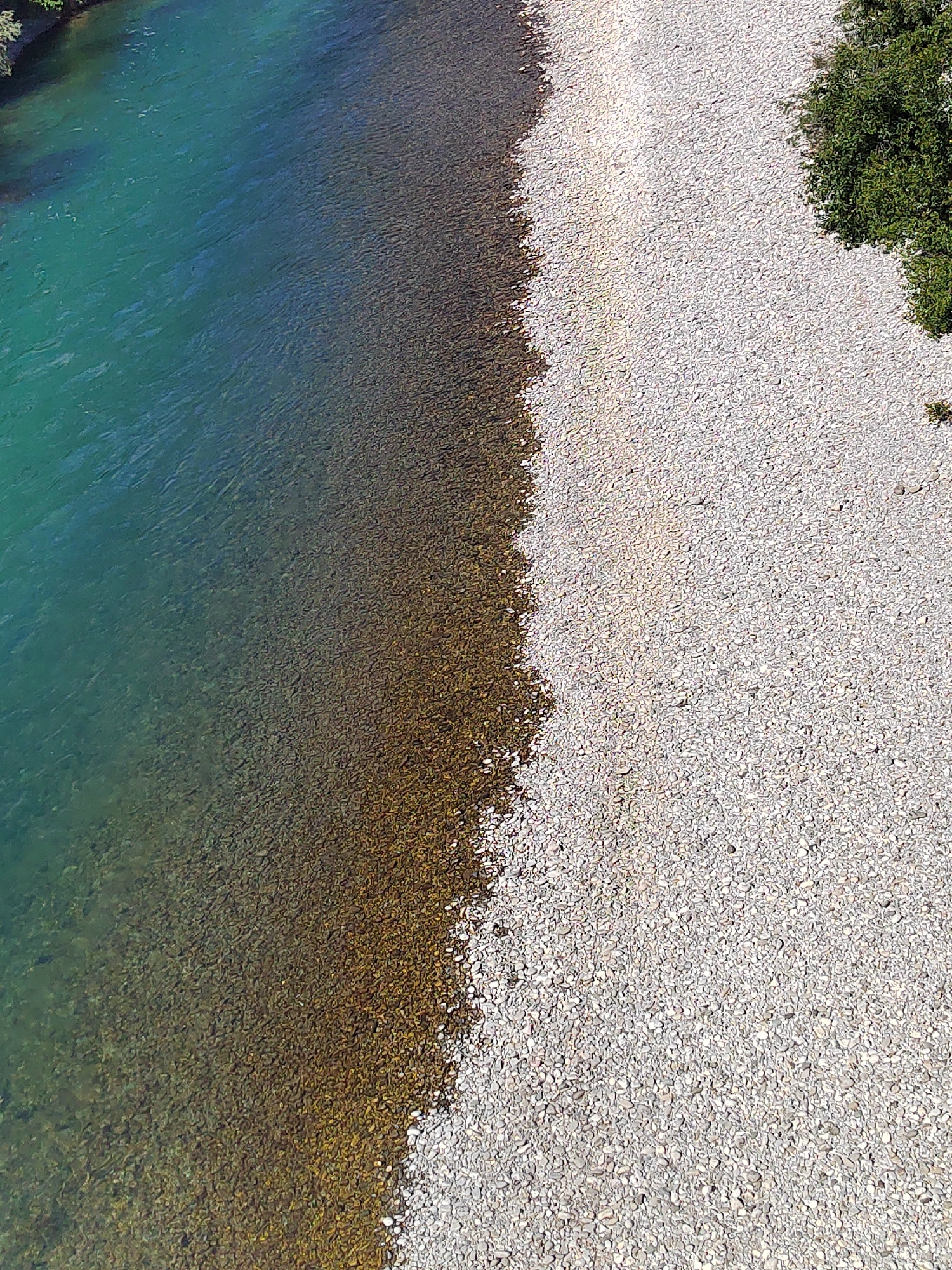 The image shows an aerial view of the Morača River's edge in Podgorica, Montenegro. The left side of the image is dominated by the river's turquoise water, which transitions to a darker, submerged rocky riverbed closer to the shore. The right side of the image features a gravel bank composed of light gray stones, with a patch of green vegetation near the top right corner.