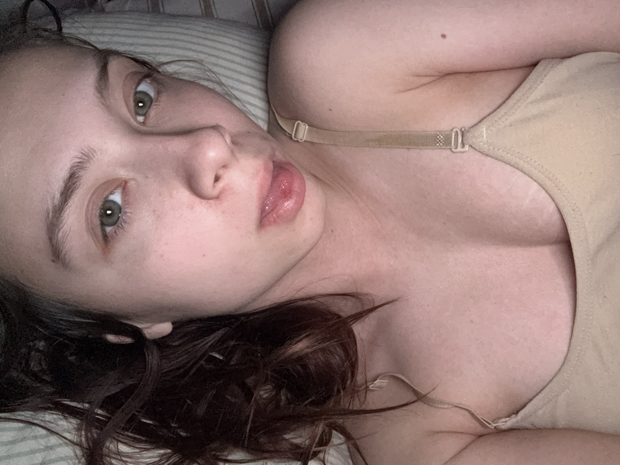 A selfie-style photograph depicts a woman lying on her back, presumably in bed. Her head is tilted towards the left, and she gazes directly at the camera. She has light skin, dark hair, and green eyes. Her lips are slightly parted, with her tongue visible. She is wearing a beige-colored tank top with a visible bra strap. The background shows a striped pillow. No information suggests the location is Прикамский, Russia.