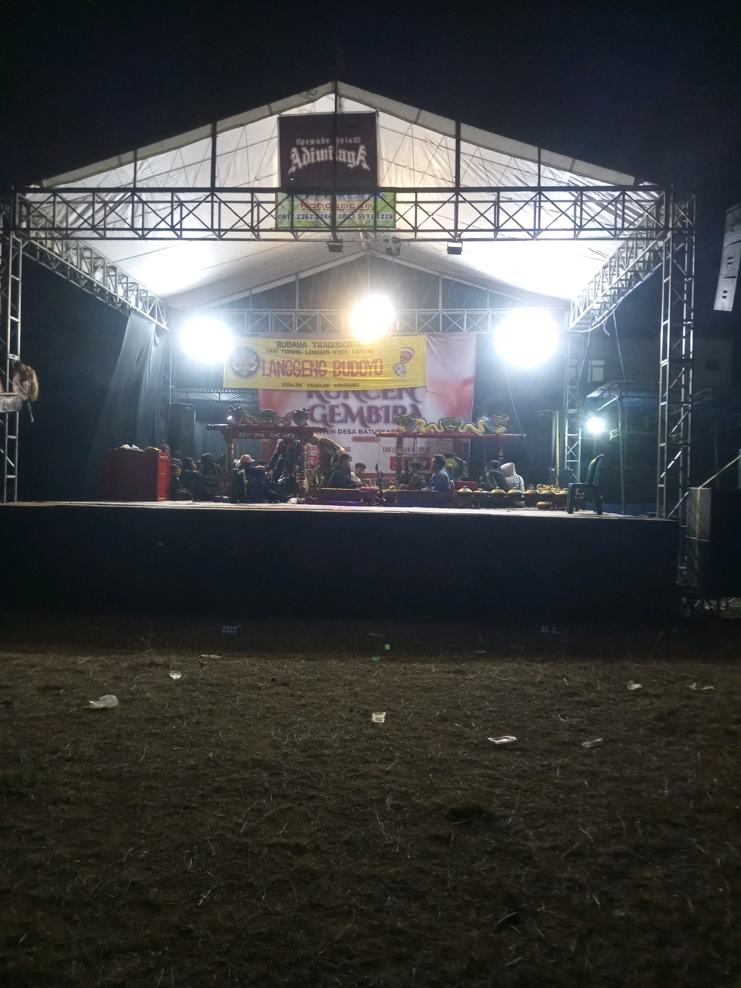 The image shows an outdoor stage set up at night in Ngadikerso, Indonesia. The stage is covered by a large tent supported by metal scaffolding. There are bright lights illuminating the stage, where a group of people are gathered, some appearing to be musicians playing traditional instruments. A banner is visible behind them with text that includes "BUDAYA TRADISIONAL" and "LANGGENG BUDOYO." The ground in front of the stage is covered in dry grass and litter. The overall scene suggests a cultural performance or event taking place in a rural or open area.