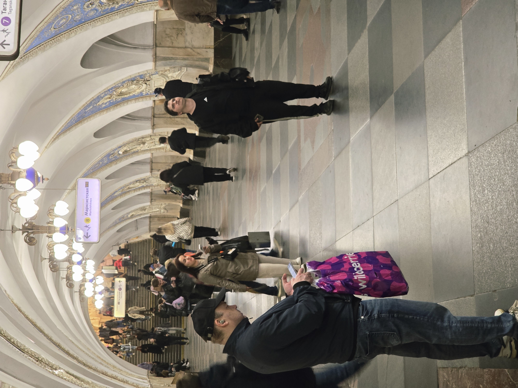 The image captures the interior of a metro station in the city of Unknown, Unknown. A group of people is walking through the station. The station has ornate architecture with arched ceilings decorated in blue and white patterns. Chandeliers hang from the ceiling, illuminating the scene. Signage, including one indicating directions to Taganskaya, is visible. The floor has a checkered pattern. A man in jeans and a jacket is looking at his phone. Another man dressed in a black tracksuit is walking through the station.