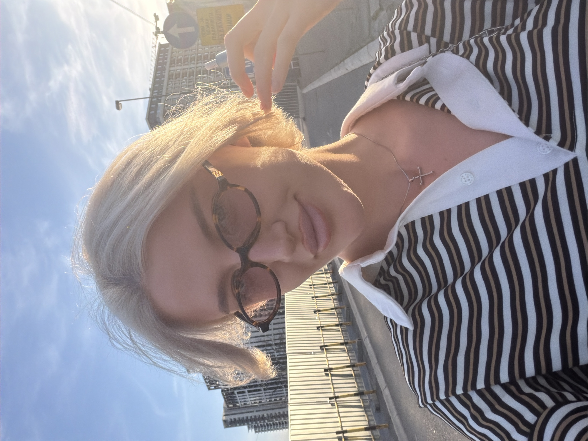 A young woman takes a selfie outdoors on a sunny day in Belgrade, Serbia. The sky is blue. She has blonde hair and is wearing sunglasses, a striped shirt with a white collar, and a silver necklace with a cross pendant. She is holding a small blue object near her hair. In the background are buildings, a street, and street signs. The city's name is not visible on the signs.