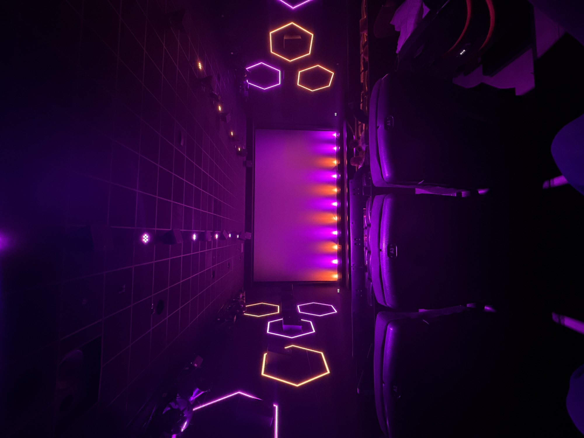 The image displays the interior of a cinema hall, situated in Kassel, Germany. The perspective is from a seated position within the audience, directed towards the front screen. The environment is illuminated by artificial light, primarily in shades of purple, pink, and orange.
The central feature is a large, blank rectangular cinema screen at the front. Below the screen, a series of individual light sources project alternating purple/pink and orange light upwards onto the screen's base. The side walls are dark and feature multiple decorative hexagonal light fixtures, some emitting orange light and others purple/pink light. The ceiling is a dark grid structure, equipped with various recessed and surface-mounted light fixtures that contribute to the purple ambient lighting.
Multiple rows of dark-colored cinema seats are visible, extending towards the screen. The headrests of the seats in the immediate foreground are labeled "A2", "A3", and "A4". These front-row seats appear to be unoccupied. In the bottom left and right corners of the frame, parts of a person's legs, clad in blue fabric, are visible, indicating the presence of the photographer. A folded white object, consistent with paper or cloth, rests on a seat or armrest in the lower right portion of the image. No other individuals are distinctly visible within the scene.