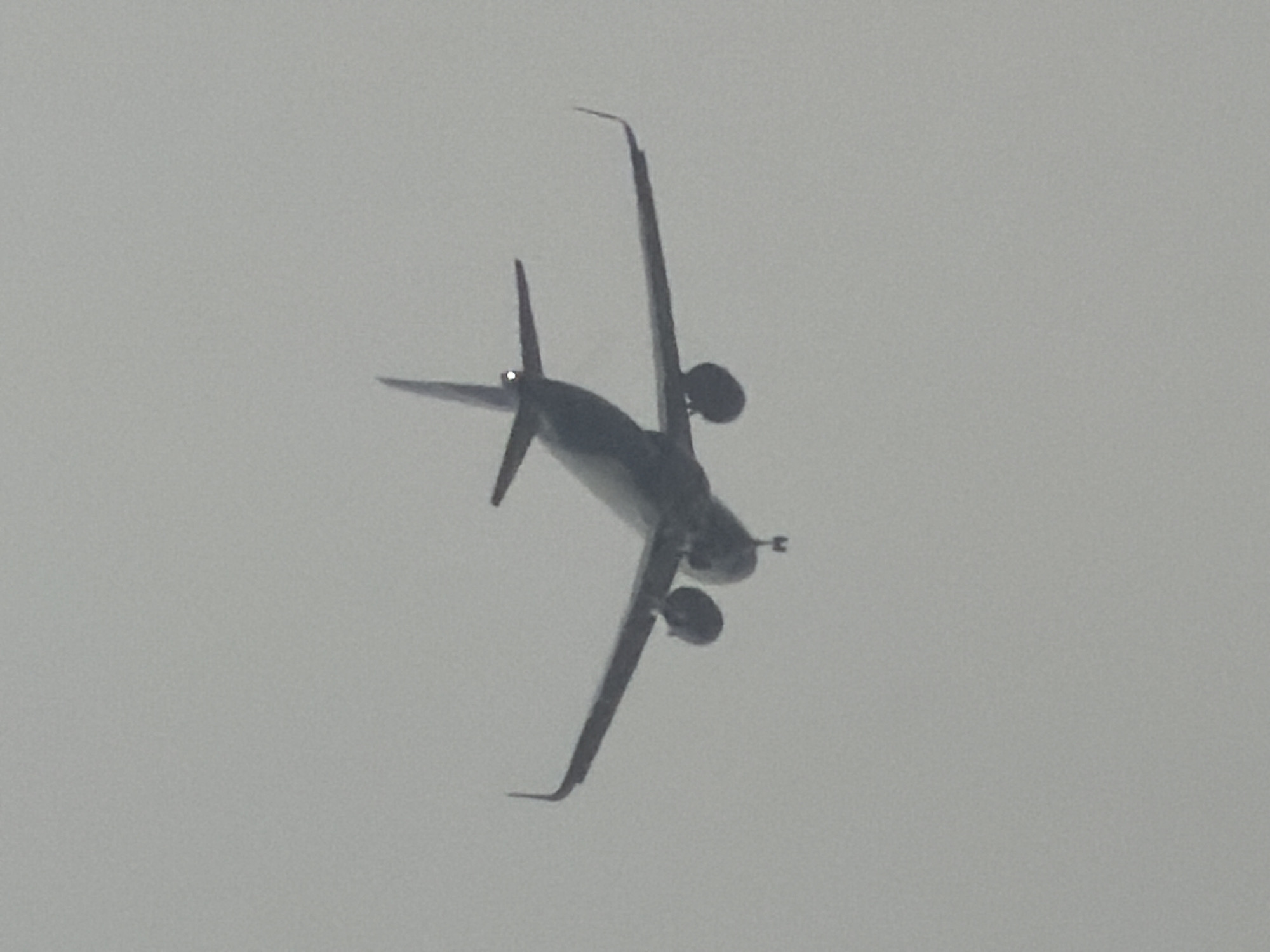 The image displays a twin-engine propeller aircraft in flight, captured from a low-angle perspective against a uniformly light grey and overcast sky. The aircraft, possibly an ATR or similar model, is seen with its underbelly facing the viewer. Prominent features include its two engine nacelles positioned under the high wings, and distinctive upward-curving winglets on both wingtips. The tail section, comprising horizontal and vertical stabilizers, is also clearly visible, with a small bright spot, possibly a navigation light, present near the base of the vertical stabilizer. The light grey background suggests cloudy, hazy, or foggy weather conditions, making it difficult to discern specific details of the aircraft's livery or to determine the precise time of day, though it is likely daytime. There are no visible people, other objects, or textual elements within the frame. The event depicted is simply an aircraft in transit. The location context for this flight is Delhi, India.