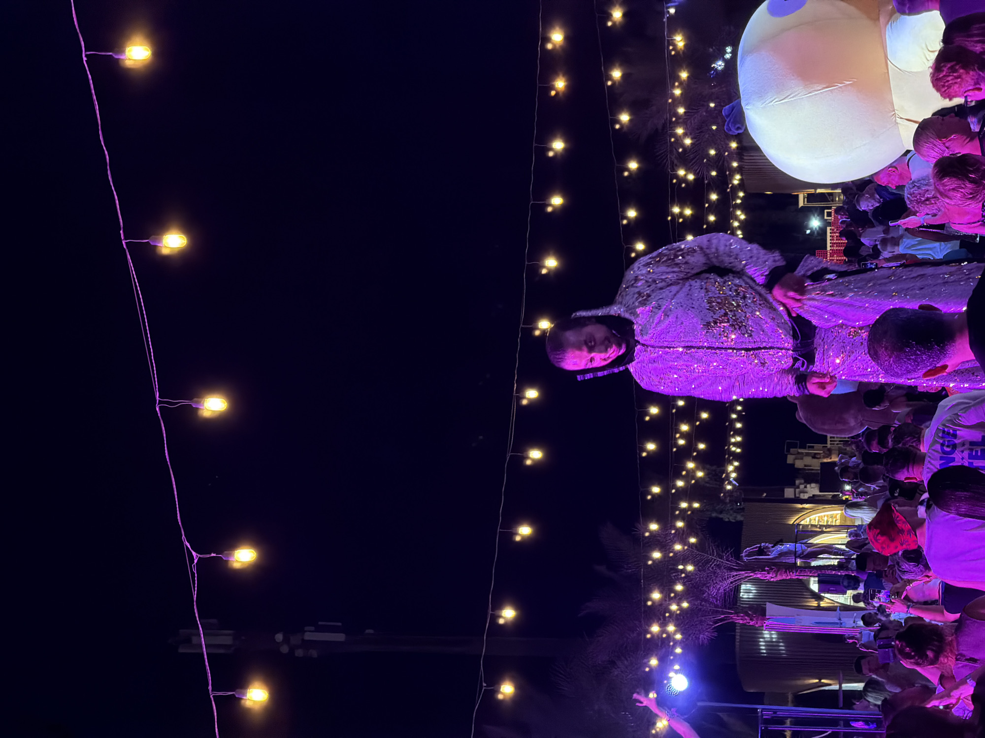 The image depicts an outdoor nighttime event, likely a festival or party, in Sharm El Sheikh, Egypt. A person in a sparkling, sequined costume stands on elevated platforms, towering over a crowd of onlookers. The person is looking downwards and slightly to their left, with a slight smile on their face. The costume is predominantly silver with gold or bronze accents. In the foreground, the backs of several attendees are visible, some holding up their phones to capture the spectacle.
The environment is illuminated by strings of yellow fairy lights that crisscross the sky, creating a festive atmosphere. Palm trees are visible in the background, suggesting a tropical or subtropical location, consistent with Sharm El Sheikh. To the right of the main subject, a large, inflatable yellow cartoon character with purple accents is partially visible. In the background, there are structures with corrugated metal siding, possibly part of temporary event infrastructure or permanent buildings. The overall scene suggests a lively and celebratory gathering.