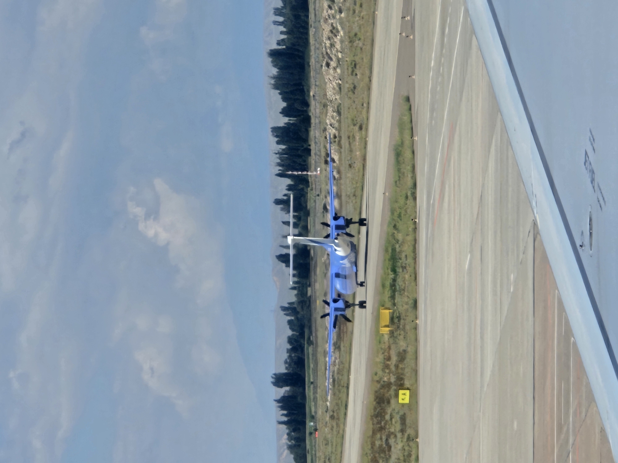 An aerial view shows a blue and white airplane on a tarmac, with a portion of another airplane's wing in the foreground. The airplane on the tarmac is a turboprop aircraft with two engines and a white tail with blue markings. It is positioned on a runway, facing away from the camera. The tarmac is grey concrete with yellow markers and painted lines. The background features a treeline and distant mountains under a cloudy sky. The scene appears to be an airport in the city of Choktal, Kyrgyzstan, given the context provided. There are no people clearly visible in the image.