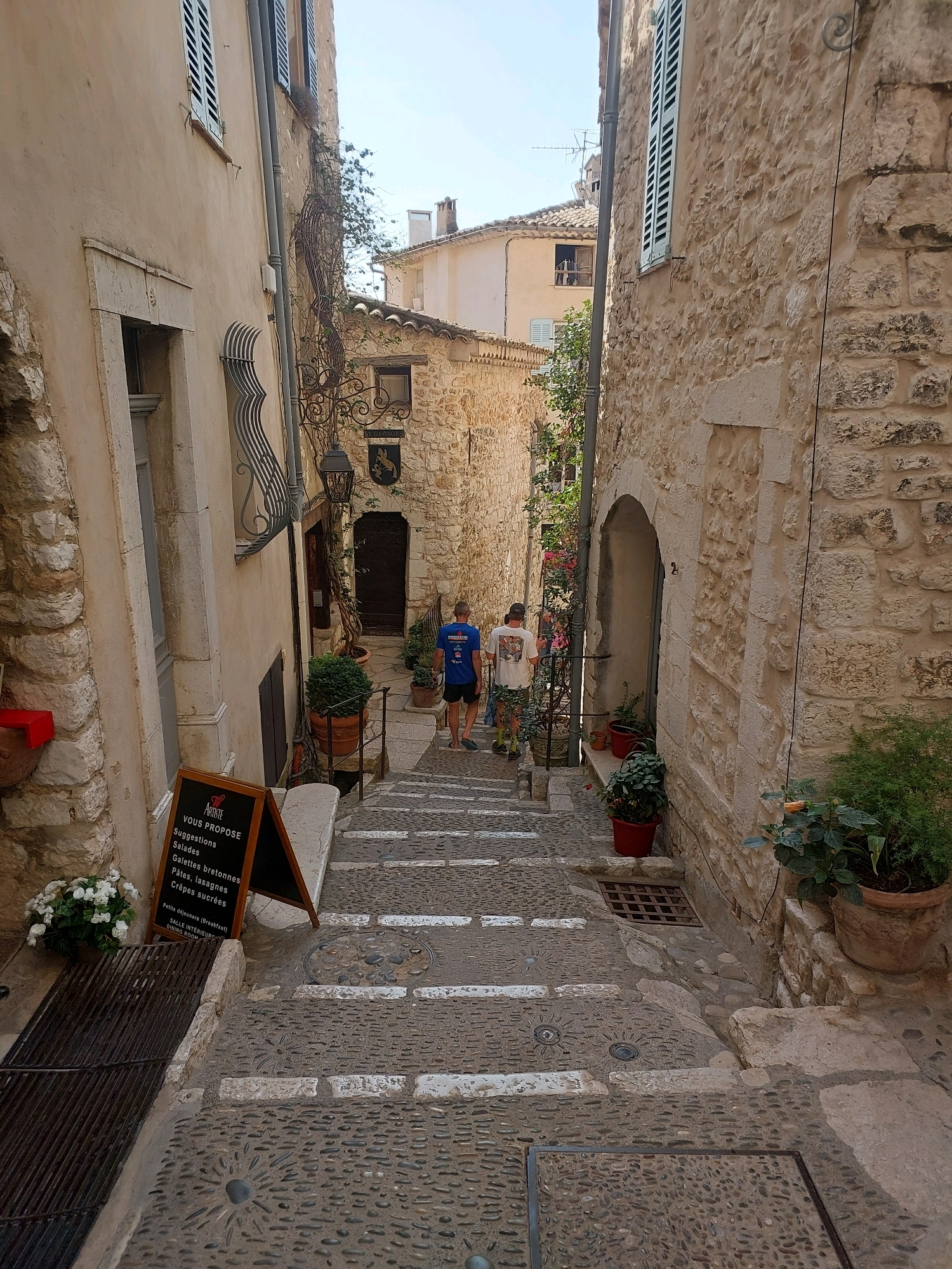 The image captures a narrow, cobblestone street in the medieval village of Saint-Paul-de-Vence, France. The street ascends steeply, lined with old stone buildings with muted, earthy tones. Two individuals are visible walking up the stairs, one in a blue t-shirt and black shorts, the other in a white t-shirt and camouflage shorts. A blackboard sign stands near the bottom left, displaying a menu in French, including "Suggestions," "Salades," "Galettes bretonnes," and "Pâtes, lasagnes." White flowers in a pot are placed to the left of the sign. The architecture features rustic stone walls, arched doorways, and wooden shutters. Potted plants and greenery are strategically placed along the walkway and on ledges, adding touches of life to the historic setting. The sky above is clear and bright. The overall scene conveys a sense of timeless charm and the picturesque character of a southern French village.
