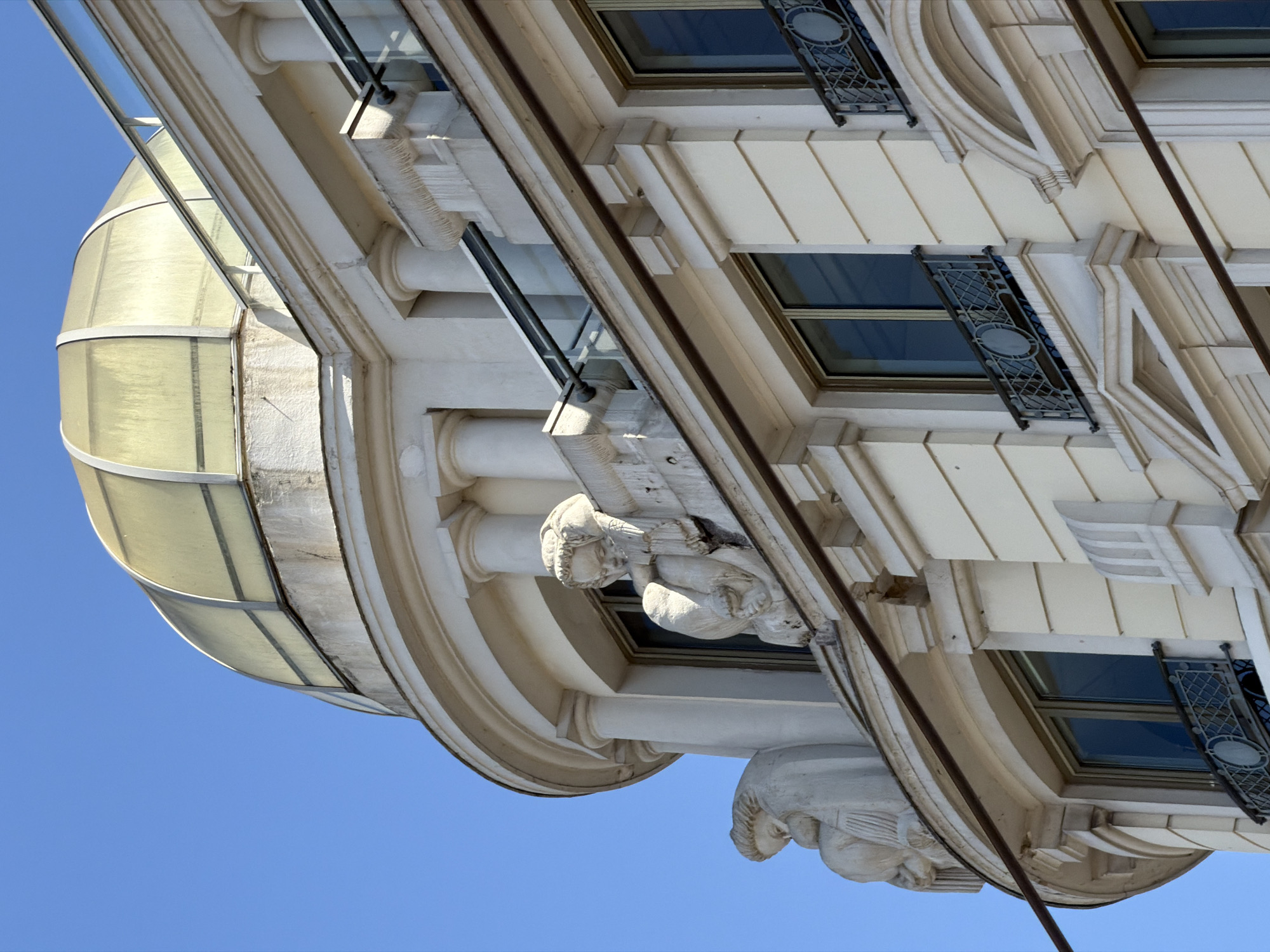 The image shows a close-up of the upper floors of a white, ornate building with a distinctive gold-tinted dome. The architecture features classical elements such as columns, decorative moldings, and sculpted figures. Specifically, there are two sculptures of child-like figures adorning the exterior of the building, one on the left leaning against a rounded corner, and another on the right seemingly peering downwards from a ledge above a window. The building has several windows with dark frames and decorative wrought-iron balconies. The sky is a clear, bright blue. The building appears to be located in Lyon, France, suggested by the architectural style often found in the city. The image is a static, daylight shot focused on the building's facade and architectural details.