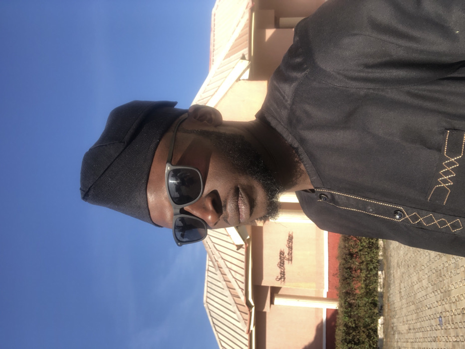 A man wears sunglasses and a dark cap, facing the camera. He has a beard and wears a black shirt with golden embroidery. The background features a building with beige walls, a brown roof, and a green hedge. The sky is a clear blue. A walkway or paved area is also visible, made of light-colored stones.