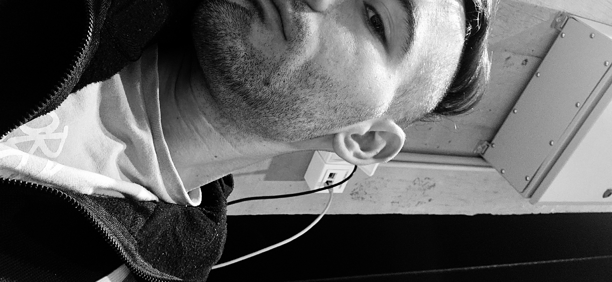 A monochrome, rotated close-up shot focuses on a man's face and upper body. The man has short dark hair, an eyebrow that is furrowed slightly, and a beard. He is wearing a shirt with a visible logo on the left side. Part of a jacket with a visible zipper is near the left side of the frame. Behind the man's head is a wall which has an electrical outlet and wiring. Adjacent to the wall, there is an unidentifiable box with visible screws. The location cannot be determined from the image.
