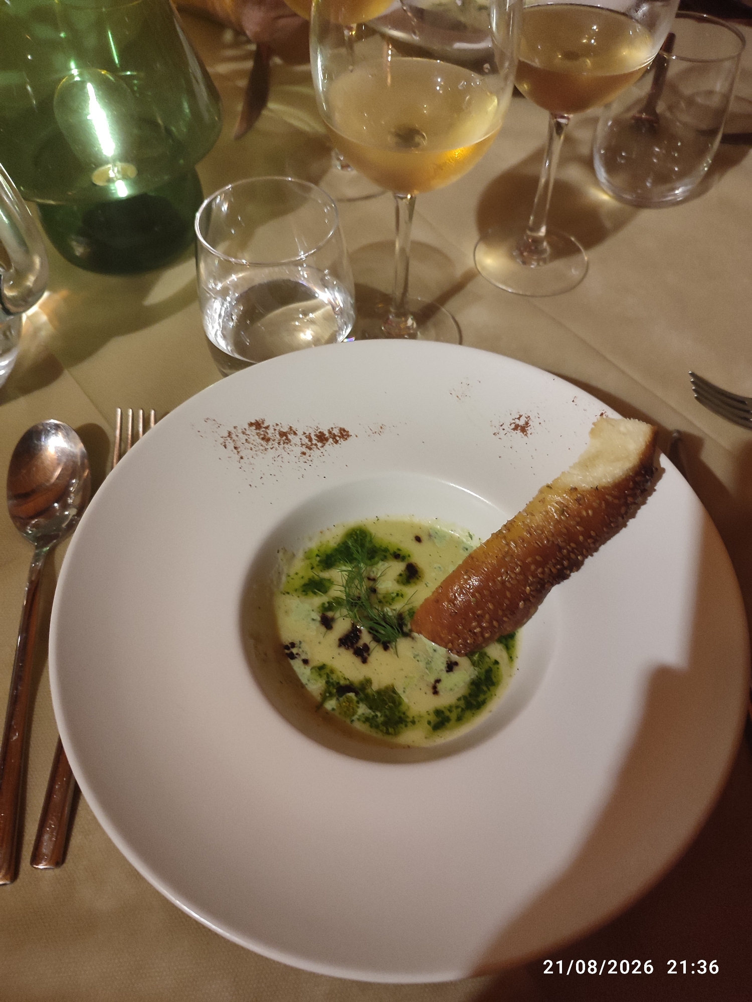 The image depicts a close-up of a white, circular plate holding a creamy soup garnished with green herbs and a piece of toasted bread. The soup appears to be a light yellow or green color, with visible pieces of herbs and possibly some dark specks. The bread, a crusty baguette slice, is placed diagonally across the soup.

The plate is set on a light brown tablecloth, which has a textured pattern. To the left of the plate, a silver spoon and fork are visible, with the spoon resting on top of the fork. In the background, several glasses are present. Two wine glasses, one partially filled with a light-colored liquid, are on the right. A smaller, clear glass with water is also visible. A green glass bottle, possibly a wine carafe, is partially visible on the far left. The overall lighting suggests an indoor setting, likely a restaurant, with warm ambient light. The timestamp "21/08/2026 21:36" is present in the bottom right corner of the image, indicating the date and time.

The scene does not provide explicit visual cues to pinpoint a specific location in Militello in Val di Catania, Italy, beyond the general impression of a dining establishment.