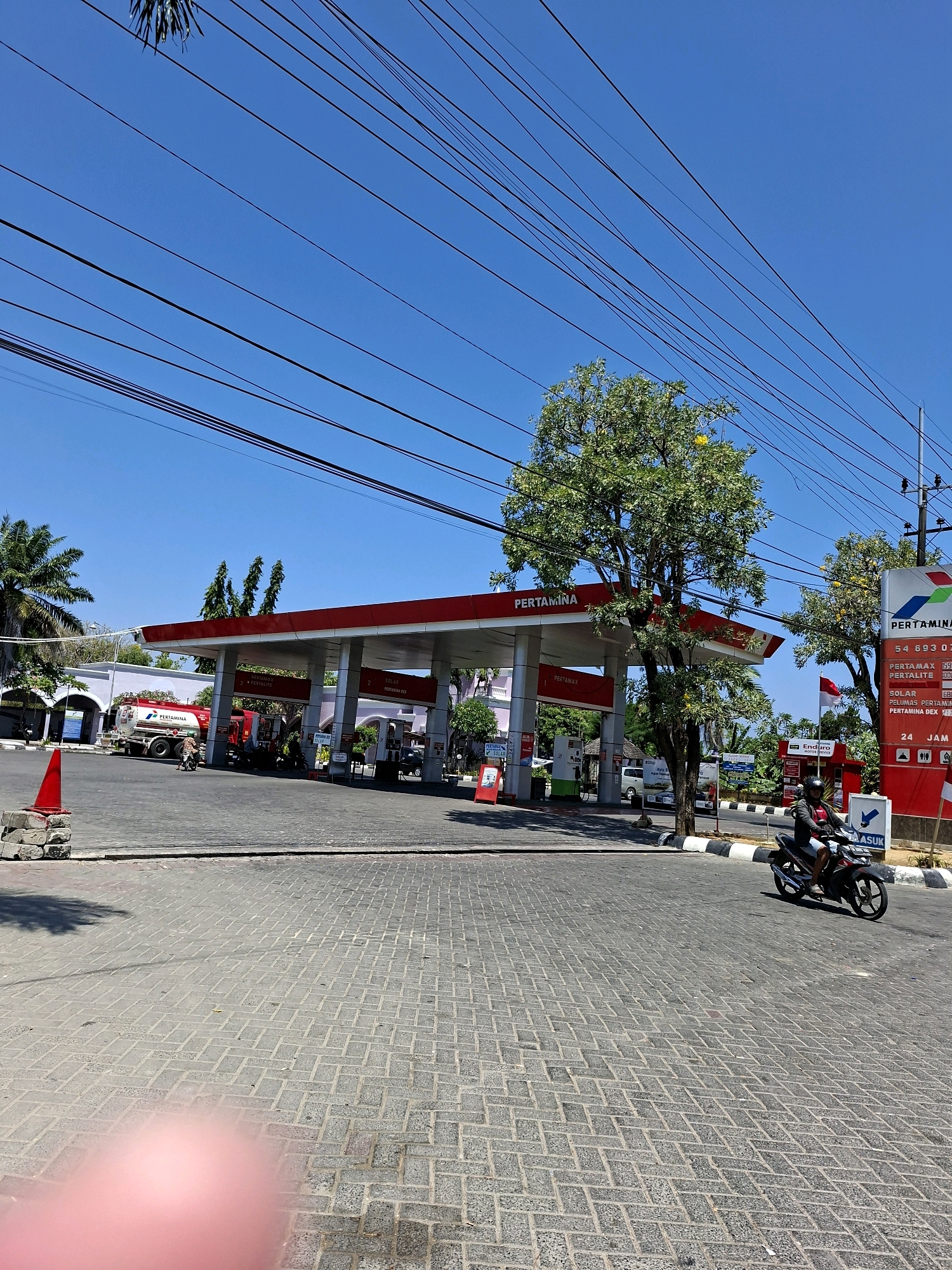 The image shows a Pertamina gas station in Larangan Tokol, Indonesia. A motorcycle is being ridden by a person in the foreground. The gas station has a red and white roof. There are several fuel pumps under the roof. A Pertamina tanker truck is parked at one of the pumps. A tall tree with green leaves is located to the right of the gas station. The sky is clear and blue. The ground in front of the gas station is paved with interlocking concrete blocks. A red traffic cone is placed on the ground to the left of the gas station. A sign with prices of fuel is visible on the right side of the image. A building is visible in the background behind the gas station. Electrical wires are strung across the sky above the gas station.