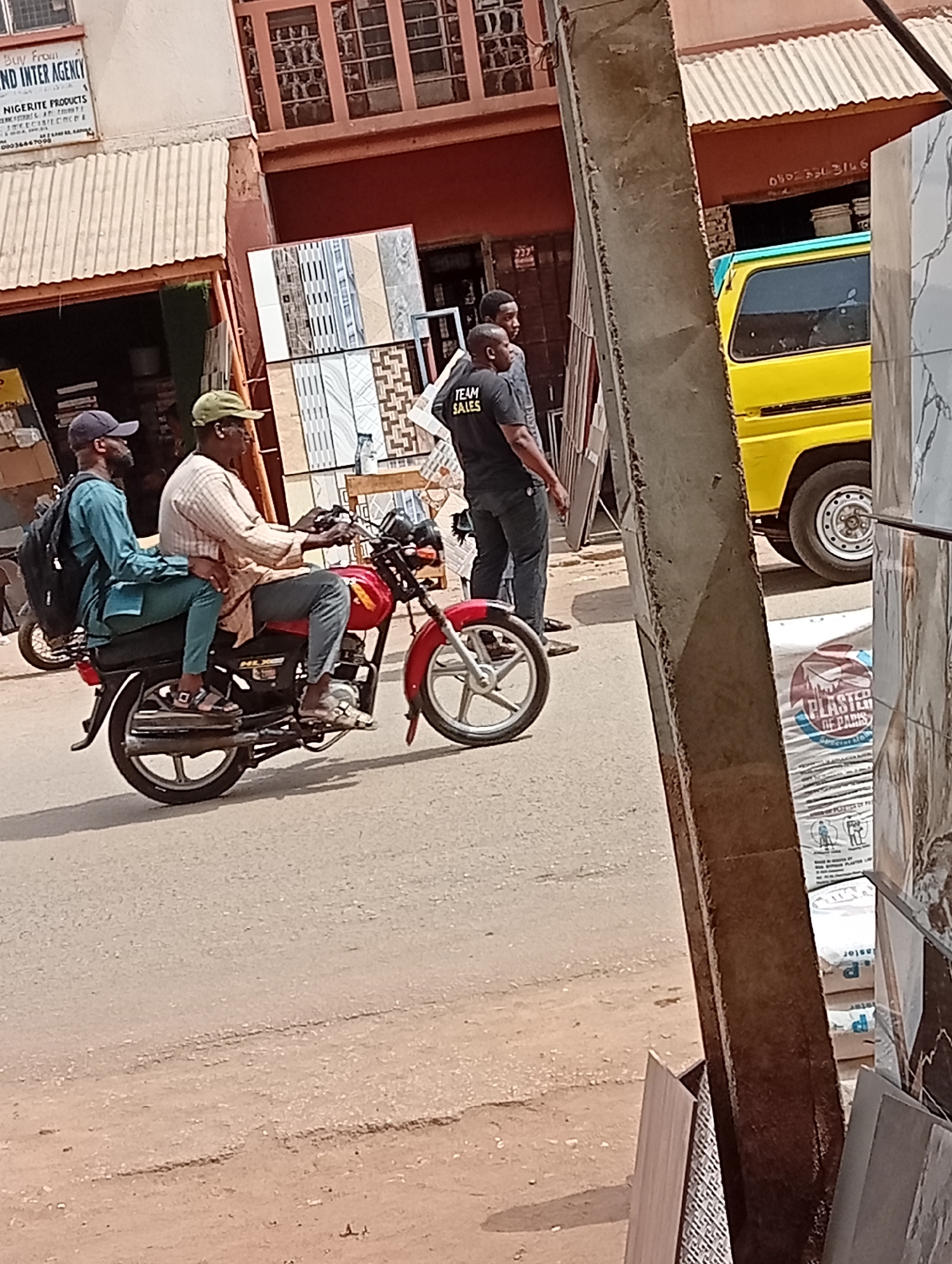 The image captures a vibrant street scene in Maiburiji, Nigeria, during daylight hours, indicated by the clear lighting and shadows. The central focus is a red motorcycle carrying two men. The driver, wearing a light-colored long-sleeved shirt and a green cap, operates the bike. Behind him, a passenger dressed in a blue-green long-sleeved garment and a dark cap sits, carrying a black backpack. Both individuals appear to be men.
On the right side of the road, two other men are standing. One wears a black t-shirt with "TEAM SALES" printed on the back, while the other stands nearby. Adjacent to them, various building materials are displayed, including stacks of decorative tiles and bags labeled "PLASTER DE PARIS". A bright yellow truck is partially visible in the background on the right.
The left side of the frame features a building with a corrugated metal awning. A sign on this building reads "Buy From ND INTER AGENCY" and mentions "NIGERITE PRODUCTS". Other visible text includes phone numbers "000360" and "08023343146", and the number "237" on another sign. The road surface appears to be asphalt with some wear and tear. The overall scene suggests a bustling commercial area, characteristic of urban life in Nigeria.