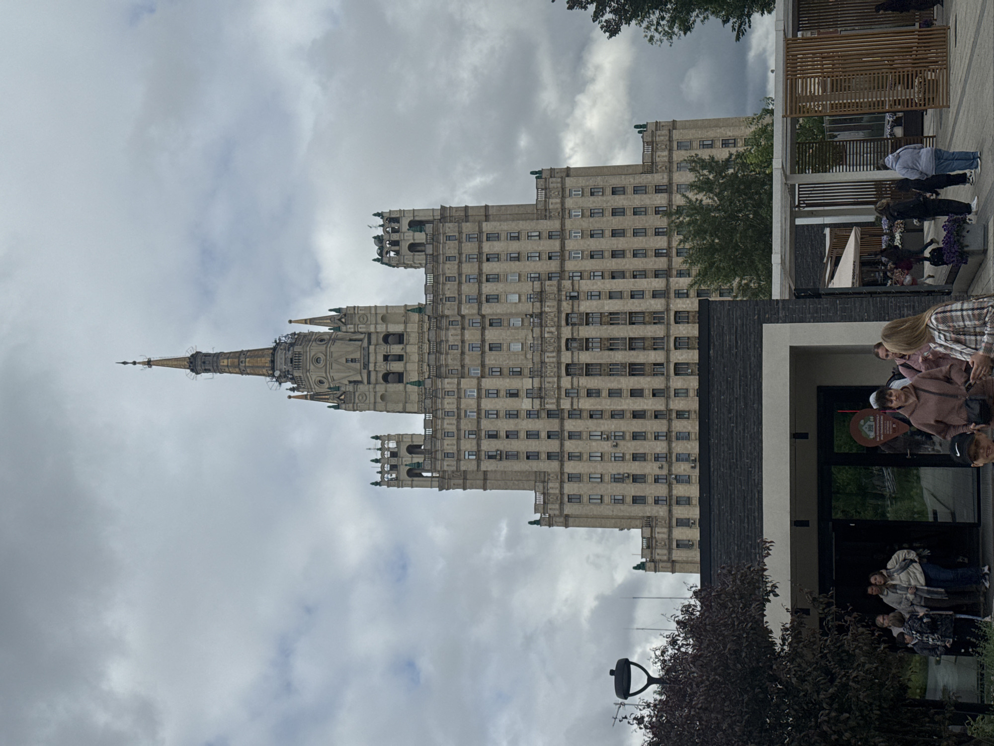 The image shows the Kotelnicheskaya Embankment Building, one of the "Seven Sisters" Stalinist skyscrapers in Moscow, Russia. The building is tall and ornate with a prominent spire. The sky is cloudy. In the foreground, there are people standing and walking near a modern building. There's also a tree and streetlamp visible.