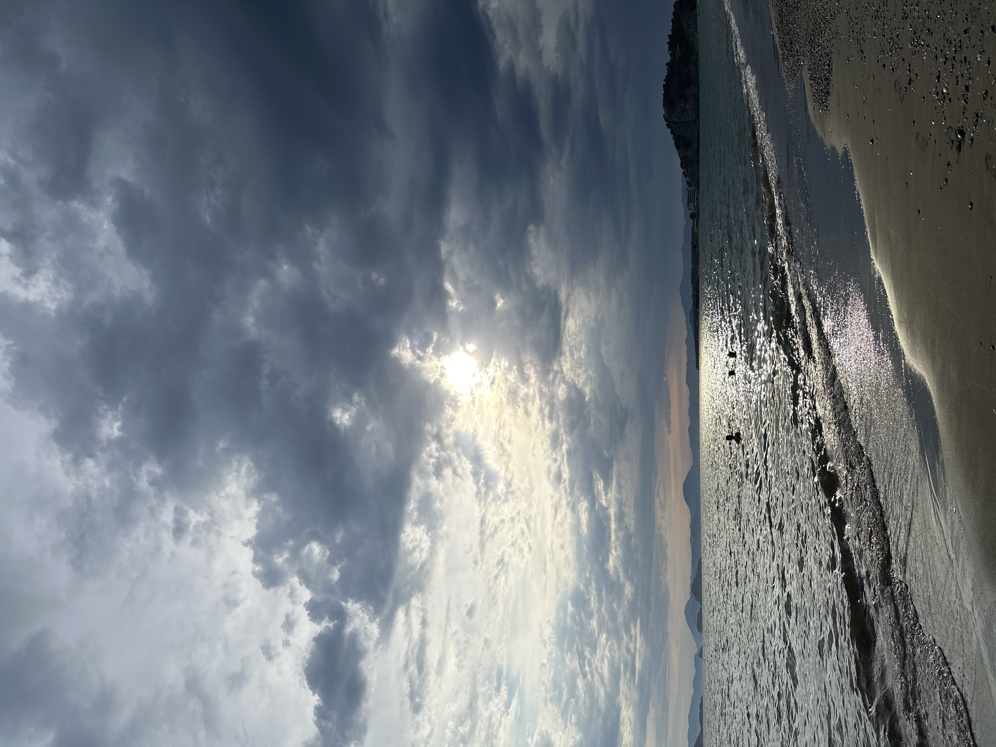 The image captures a beach scene in Fethiye, Turkey, under a dramatic sky filled with dark, heavy clouds partially obscuring the sun. The shoreline is on the right, transitioning from wet sand reflecting sunlight to dry sand. The sea extends to the horizon, reflecting the sunlight intensely. Two people are visible in the water, possibly swimming or wading. In the distance, a coastline with cliffs and distant mountain ranges is visible under the fading light. The scene is illuminated by both direct and diffused sunlight, creating a dynamic contrast between light and shadow.