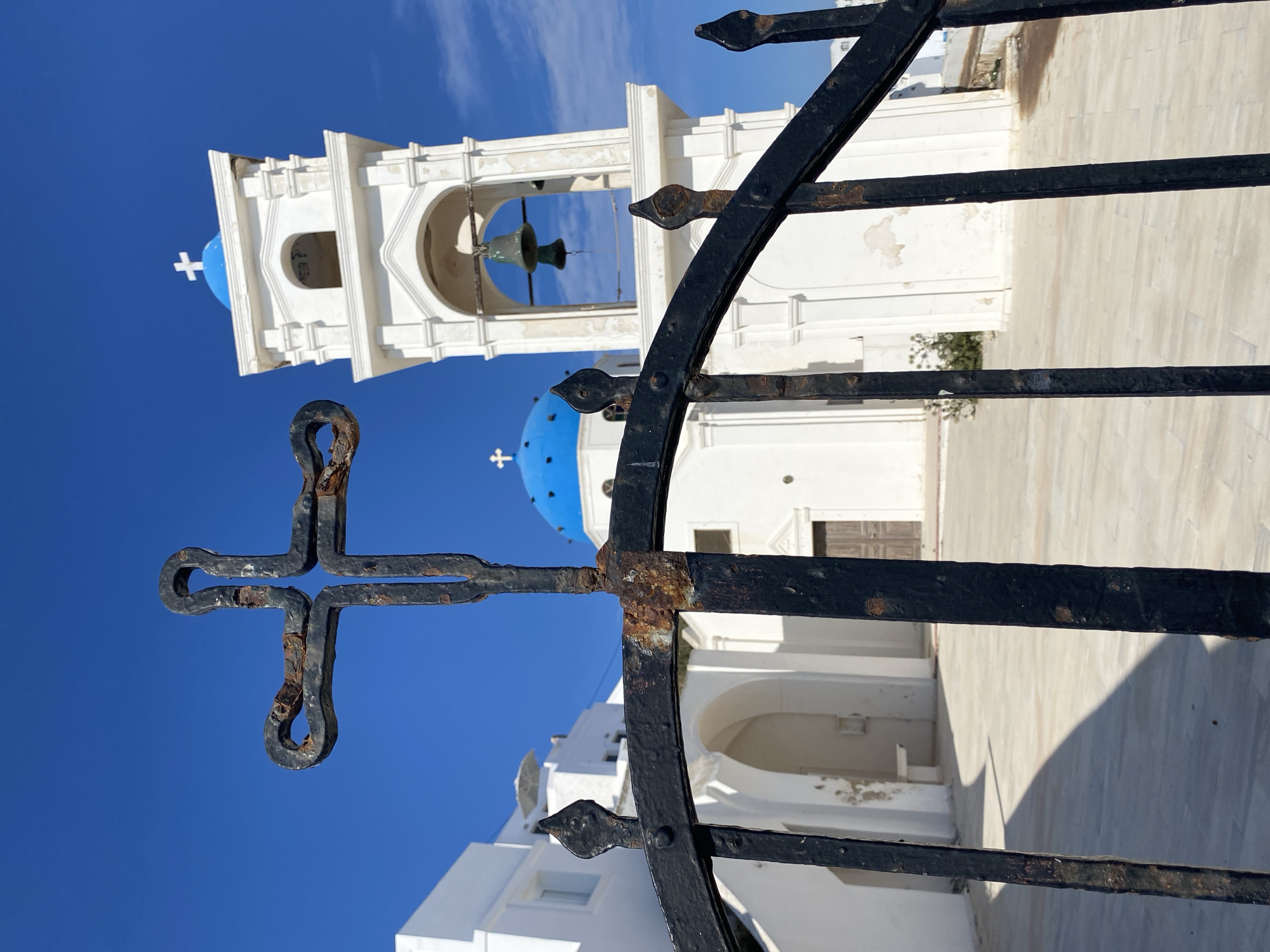 This image depicts a view of a white building with blue domes and a bell tower, likely a church, in Thira Municipal Unit, Greece. In the foreground, a rusted black wrought-iron gate with a cross motif obscures part of the scene. The sky is a clear, bright blue. The architectural style suggests a Greek island setting, possibly Santorini, with its characteristic white buildings and blue accents. There are no people visible in the image.