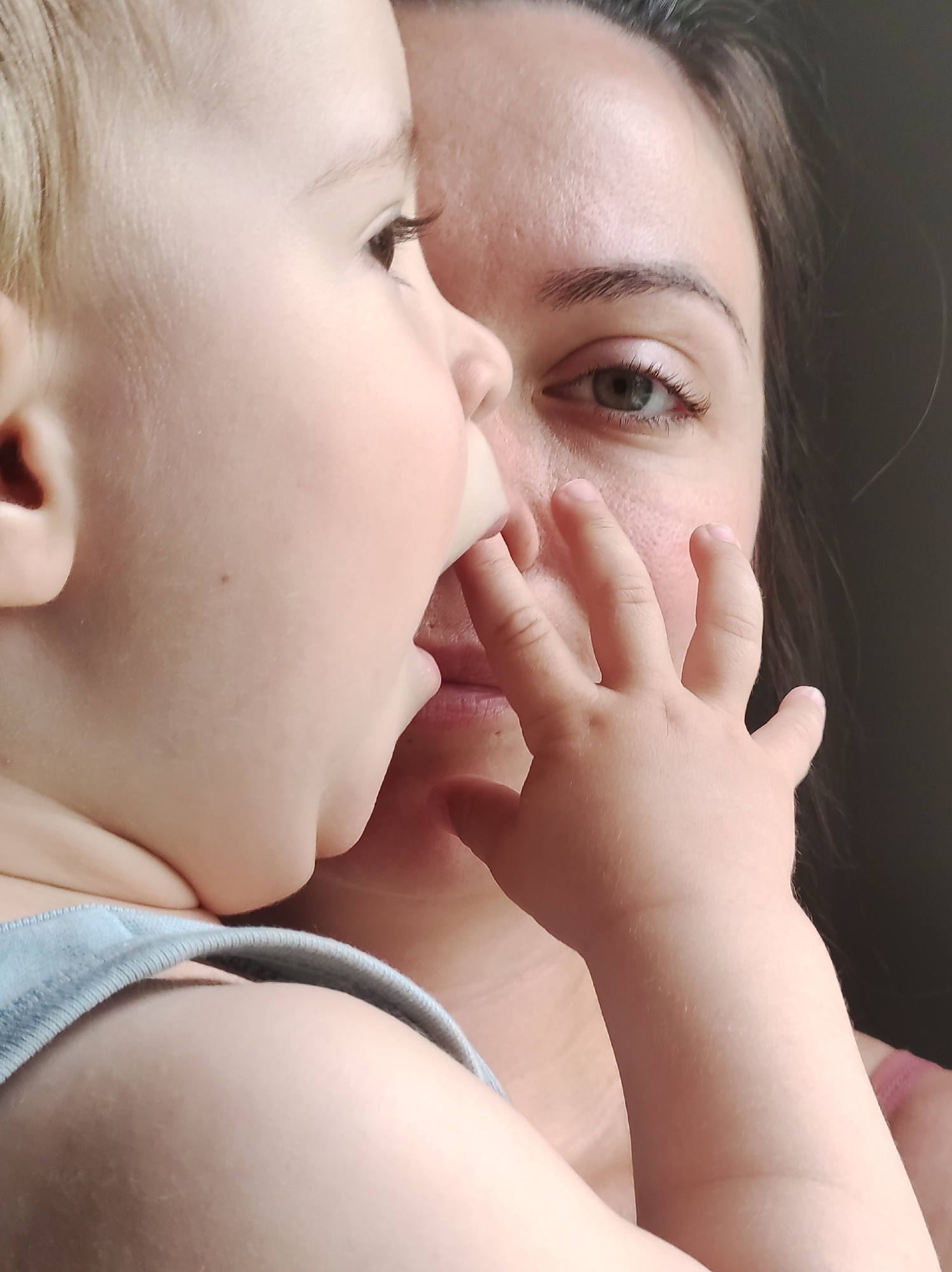 A close-up image captures a moment between a woman and a young child. The child, positioned predominantly on the left, is fair-skinned with light blonde hair and is wearing a light blue garment. The child's hand is raised and appears to be touching the woman's face. The woman's face is partially visible behind the child, revealing an eye and part of her face. The background is dark and out of focus. The photograph seems to be taken indoors, and the details of a city in Russia (Фоки) are not visually discernible from the image.