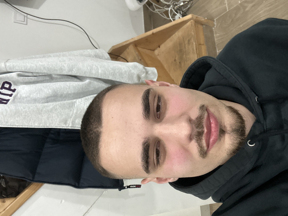 The image features a young adult male positioned centrally, captured in a selfie format within an indoor residential setting in Bragança, Portugal. The man has a short, buzzed hairstyle and dark facial hair consisting of a mustache and a small goatee. His eyes are partially closed or directed downwards, and his lips are slightly parted. He is wearing a dark, possibly black, hooded sweatshirt.
In the background, to the upper left, a dark blue padded jacket hangs against a white wall. To the upper right, a light grey hooded sweatshirt with dark lettering, possibly "IP," hangs. Below the grey sweatshirt, a light wooden staircase with visible steps ascends to the right. A white electrical outlet is mounted on the wall above the stairs. On the floor to the right, next to the base of the stairs, is a tangle of white and black cables and wires, alongside a white object resembling a small appliance or housing. The floor appears to be covered with light-colored tiles. The walls are uniformly white, and the lighting is consistent with interior artificial illumination.