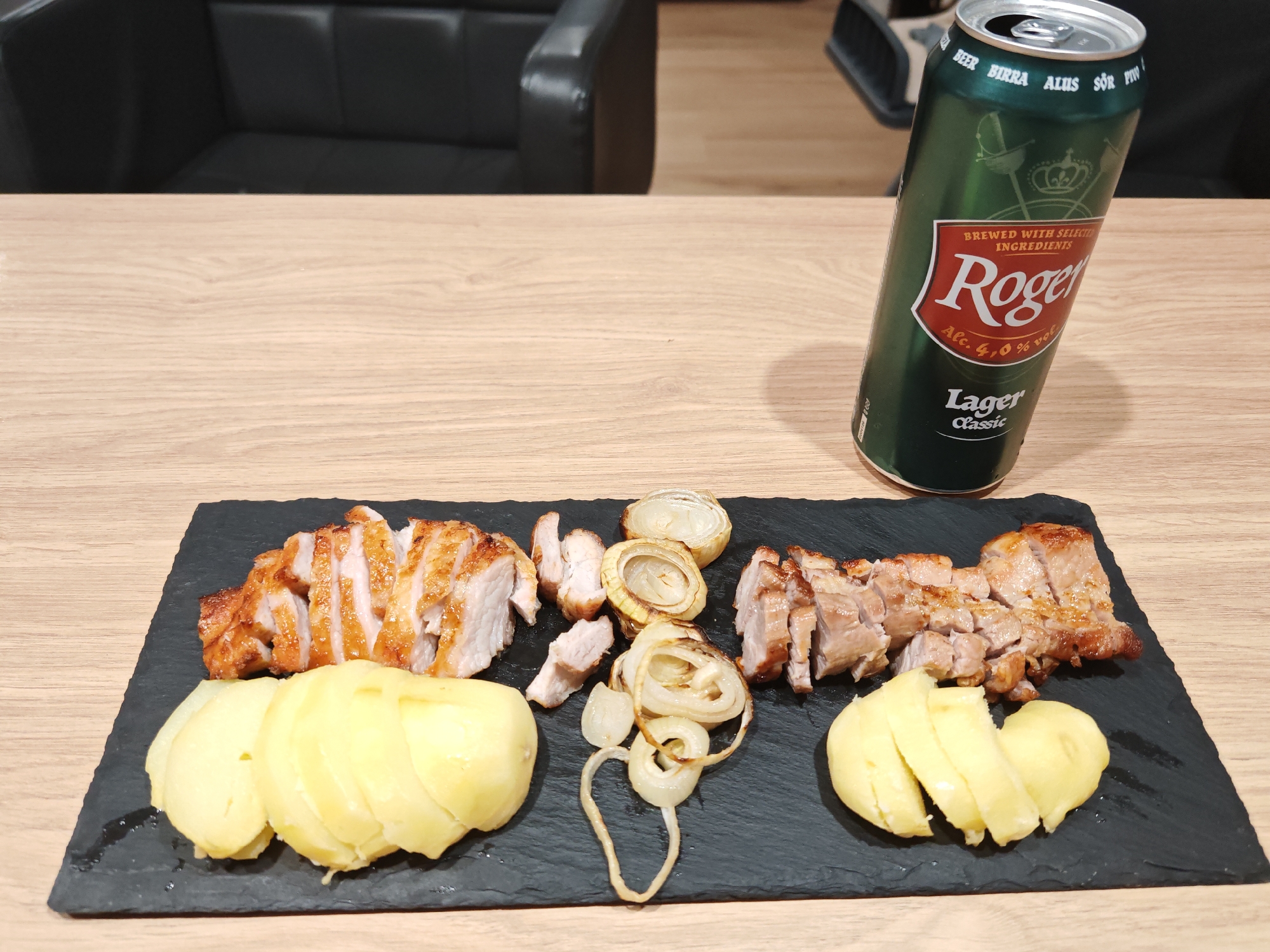 A platter of sliced roasted pork and boiled potatoes rests on a dark slate surface. The pork is arranged in two sections, one larger on the left and one smaller on the right, with some loose pieces scattered between them. Sliced grilled onions are interspersed with the pork. Two portions of boiled potatoes, each appearing to be a whole potato cut in half, are placed to the left and right of the meat. A green can of "Roger Lager Classic" beer, tilted and partially obscured, stands to the upper right of the platter. The food and drink are set on a light-colored wooden table. In the background, a portion of a dark leather chair or sofa is visible. The scene is indoors, likely in a dining area. The provided location of Plovdiv, Bulgaria is not visually verifiable from the image.