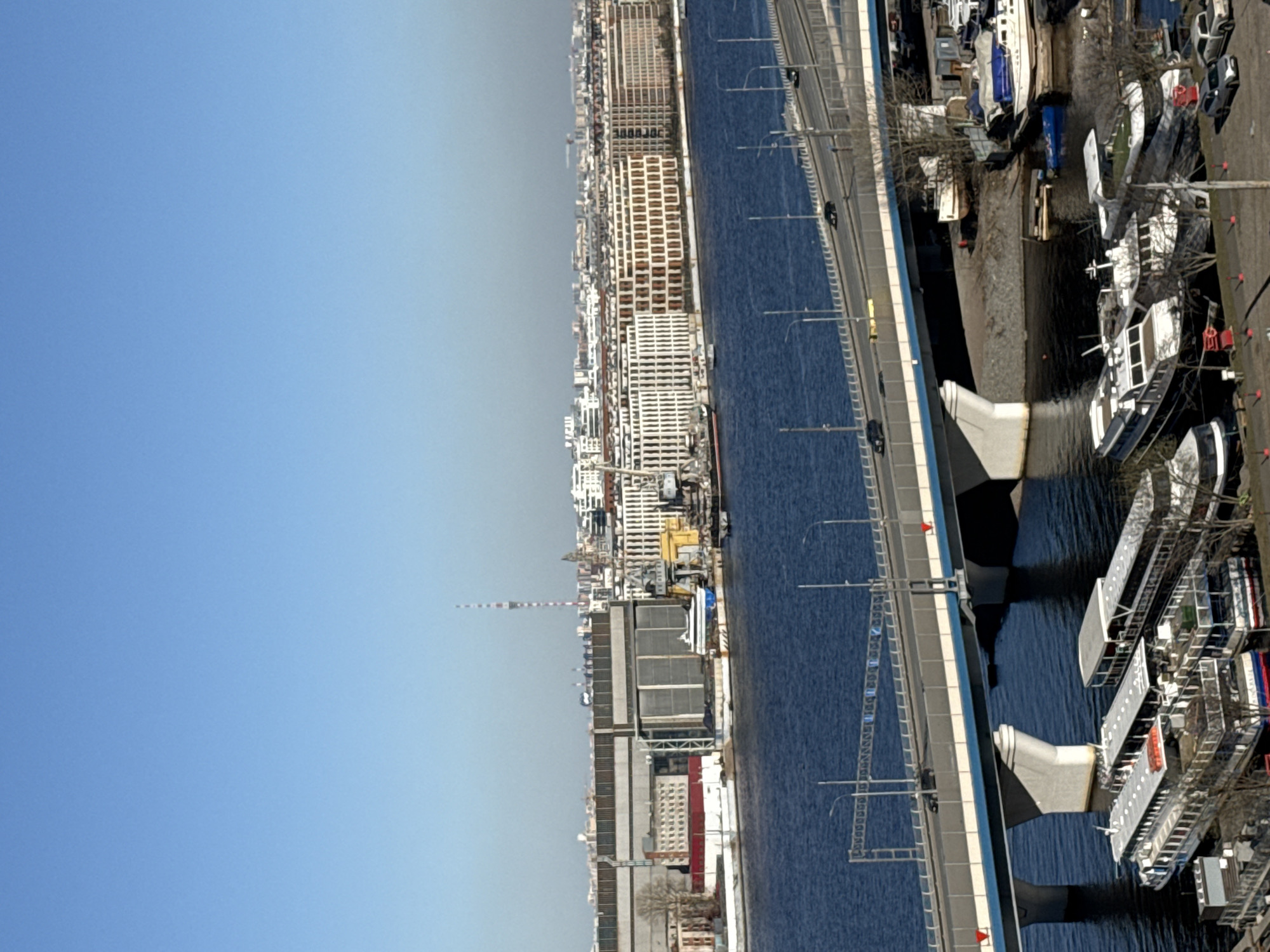 The image shows a high-angle view of a waterway flowing through a cityscape, likely in Saint Petersburg, Russia. A bridge spans the waterway, with cars moving across it. Numerous boats and ships are docked along the bank. The buildings line up along the shore with a mix of modern and older architecture. A tall telecommunications tower is visible, adding to the city's skyline. The sky is clear and blue.