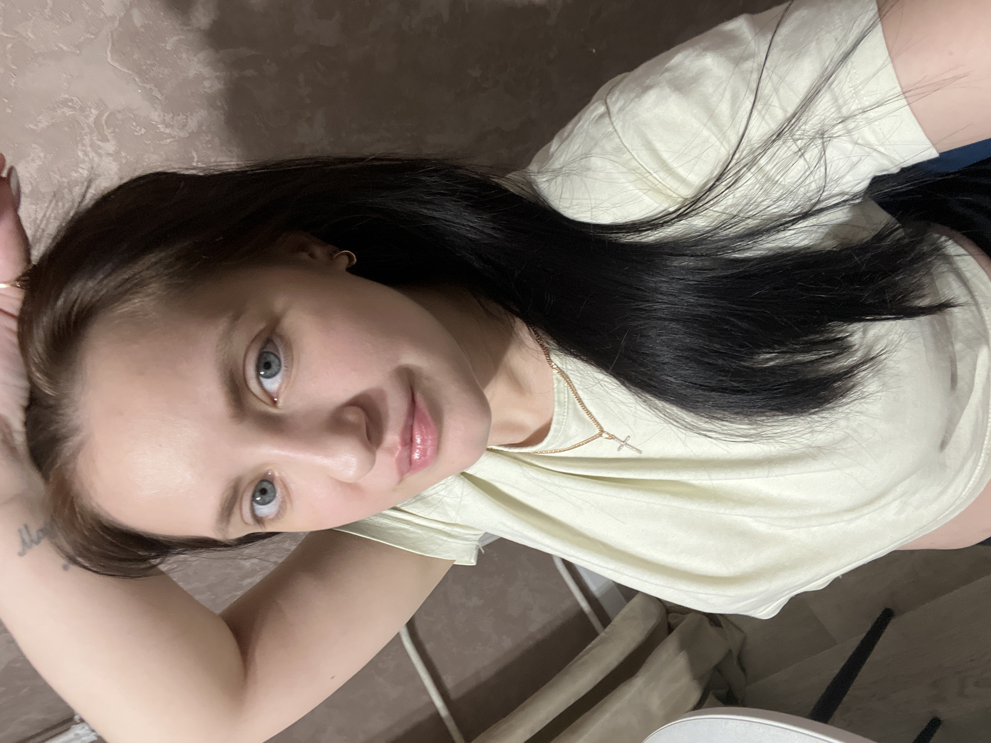 A young woman with dark brown hair and light blue eyes is posing for a close-up photo indoors. She is wearing a light-colored t-shirt and a gold necklace with a cross pendant. She is resting her right arm behind her head, partially revealing a tattoo on her inner arm. The background is a light brown wall. The photo appears to have been taken in a private residence, based on the chair visible in the bottom right corner. There is no verifiable location information that places the image in Марковский, Russia.