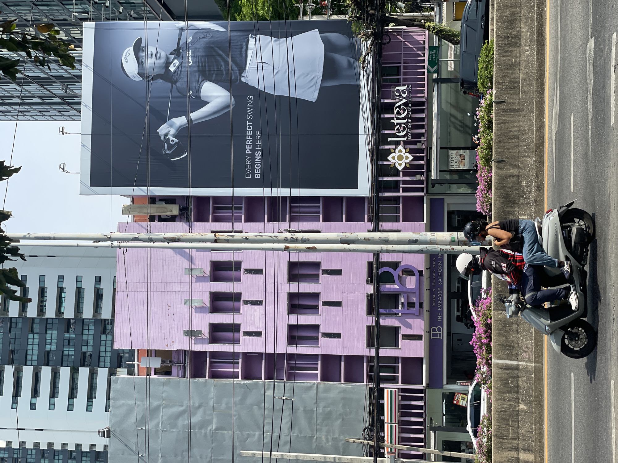 This image captures a vibrant, sunlit urban street scene, likely in Bangkok, Thailand. The central focus is a large, monochrome billboard featuring a female golfer in a cap, holding a golf club, with the text "EVERY PERFECT SWING BEGINS HERE."
Below the billboard is a distinctive purple building. The upper section of this building hosts "leteva BOUTIQUE MASSAGE & SPA," with its name in white stylized script and a floral emblem. The lower part of the building is identified as "EB THE EMBASSY SATHORN HO," with accompanying Thai script for "โรงแรม ดิ เอ็มบาสซี่ สาทร" (The Embassy Sathorn Hotel). A smaller sign outside reads "HAPPY HOUR," and various windows and architectural details are visible on the purple facade.
To the right of the purple building, a multi-lane road is visible, separated by a concrete barrier adorned with pink flowers. Two individuals on a gray scooter are riding on the road, both wearing helmets. Overhead, numerous power lines crisscross the frame, a common sight in the city. To the left, a modern building with a facade of white vertical elements and dark windows is partially visible, reflecting the bright sky. The overall impression is a bustling daytime street with prominent commercial establishments.